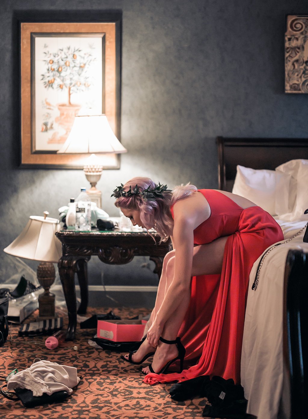 A woman in a red dress adjusts her high heels in a dimly lit room. She sits on a bed next to a bedside table with a lamp, drinks, and miscellaneous items. The room is decorated with art and has a patterned carpet.