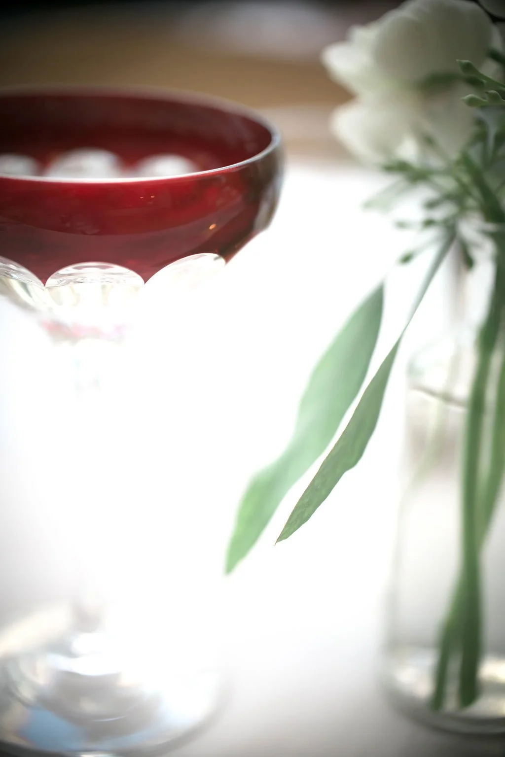 Close-up of a red glass cup with decorative patterns, next to a small vase with green leaves and white flowers, set on a white surface.