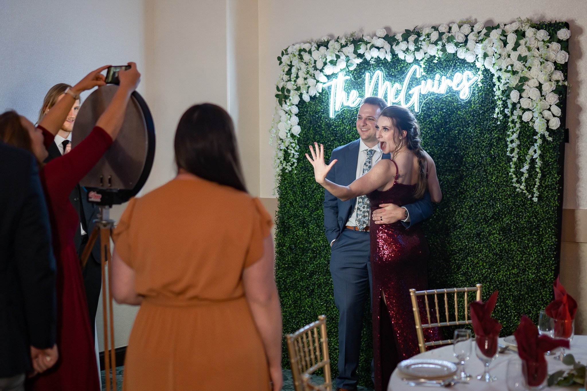 A couple posing for a photo in front of a greenery wall with "The McGuires" sign at a formal event, while someone takes their picture with a smartphone.