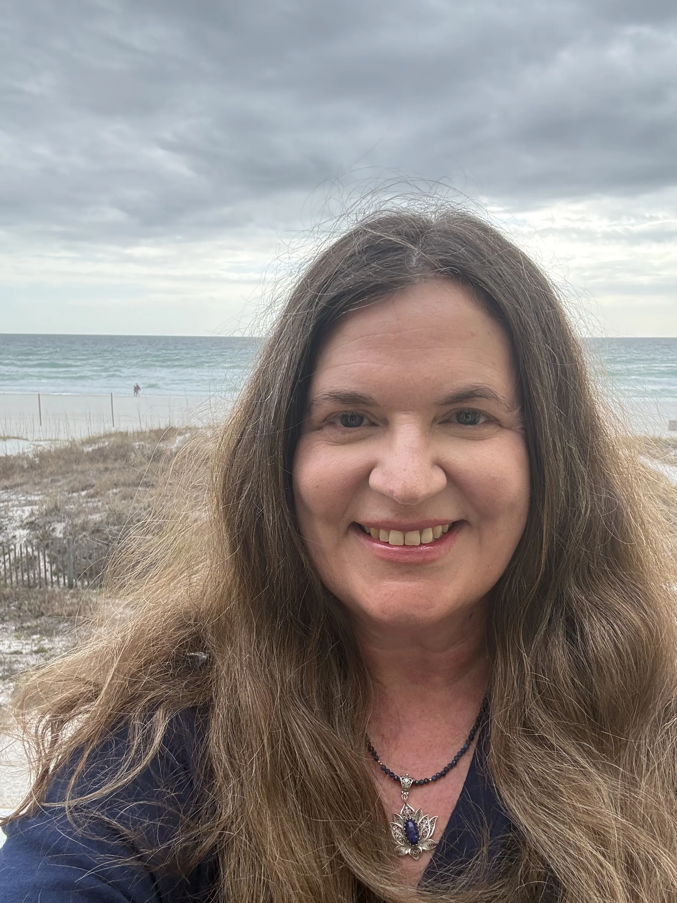 Smiling woman with long hair taking a selfie at the beach with the ocean and cloudy sky in the background.