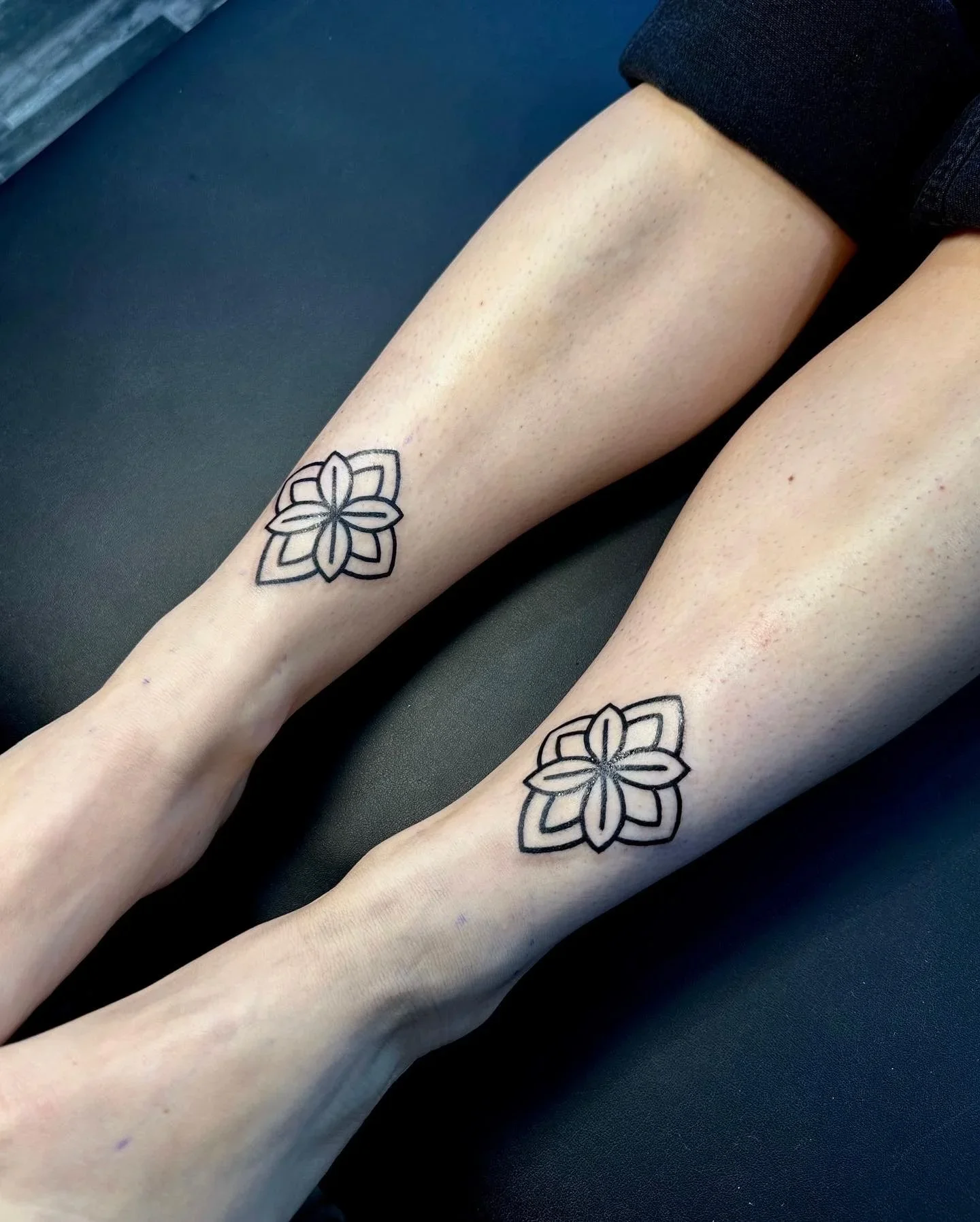 Two person's legs with matching black geometric floral tattoos on their calves, lying on a black surface.