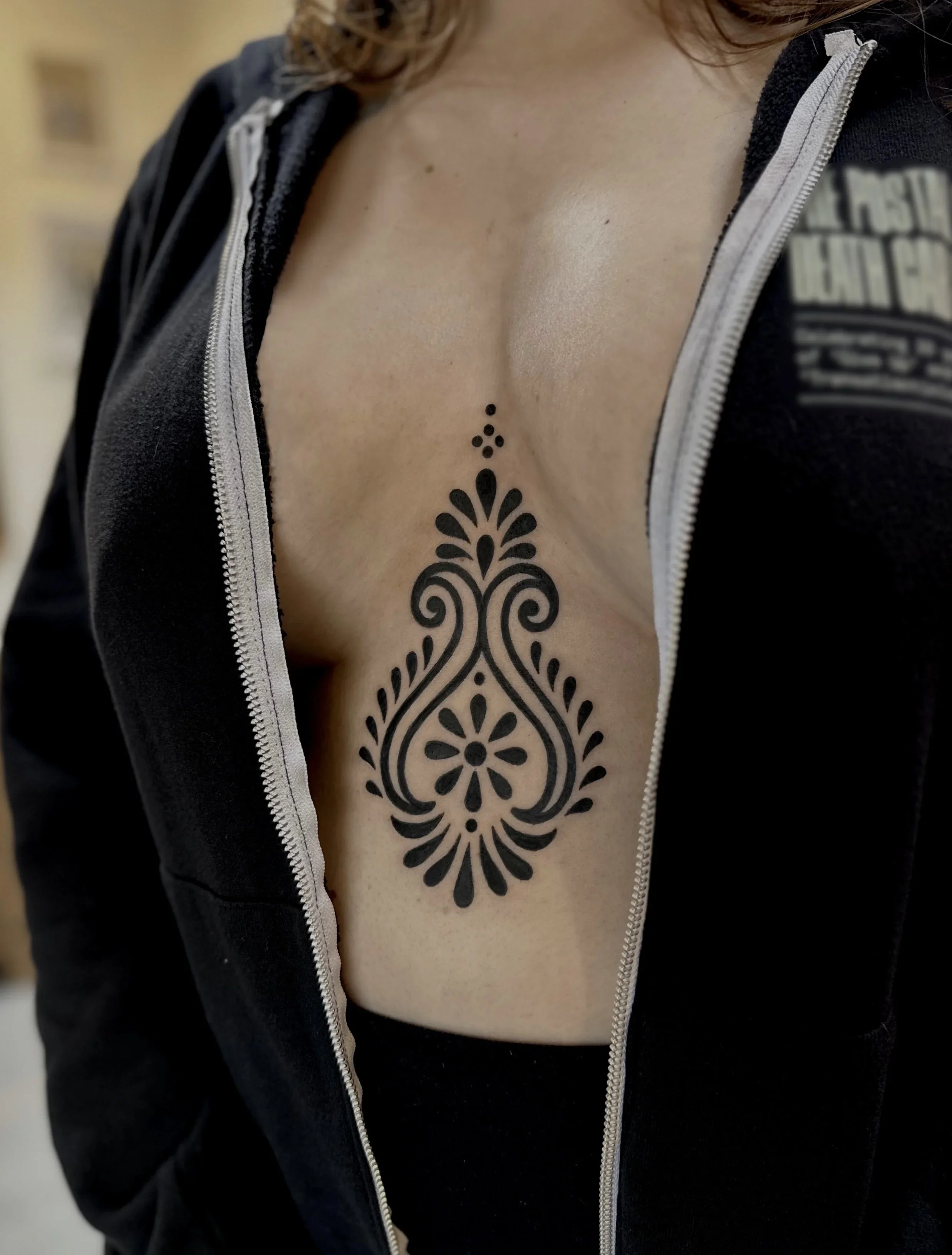 Close-up of a woman's chest showing a large black tattoo with intricate floral designs extending downward on her skin, partially covered by a black zip-up hoodie.