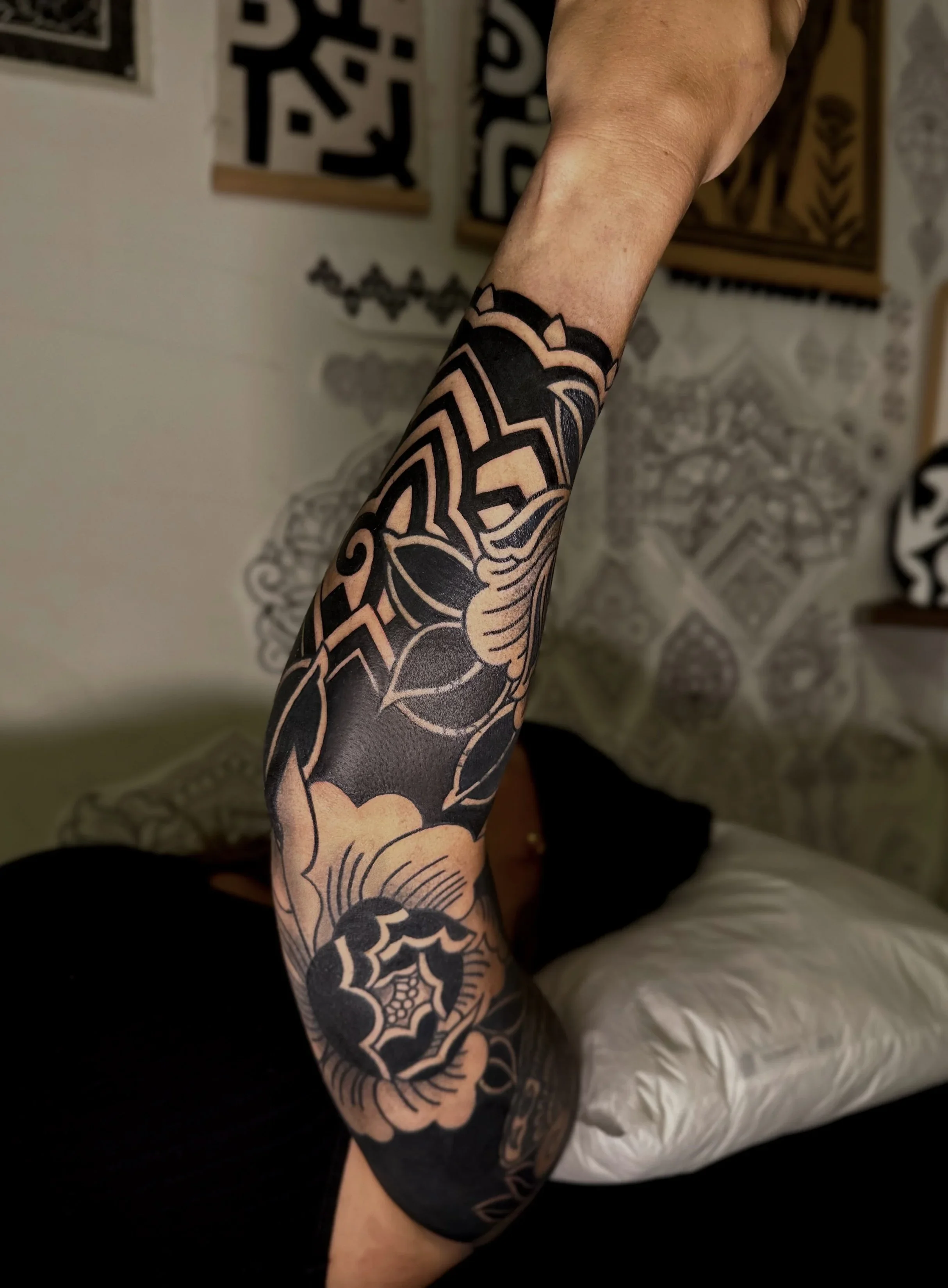 A person's arm with a black and gray tattoo sleeve that includes floral and geometric patterns. In the background, a table with a patterned tablecloth and framed artwork on the wall are visible.