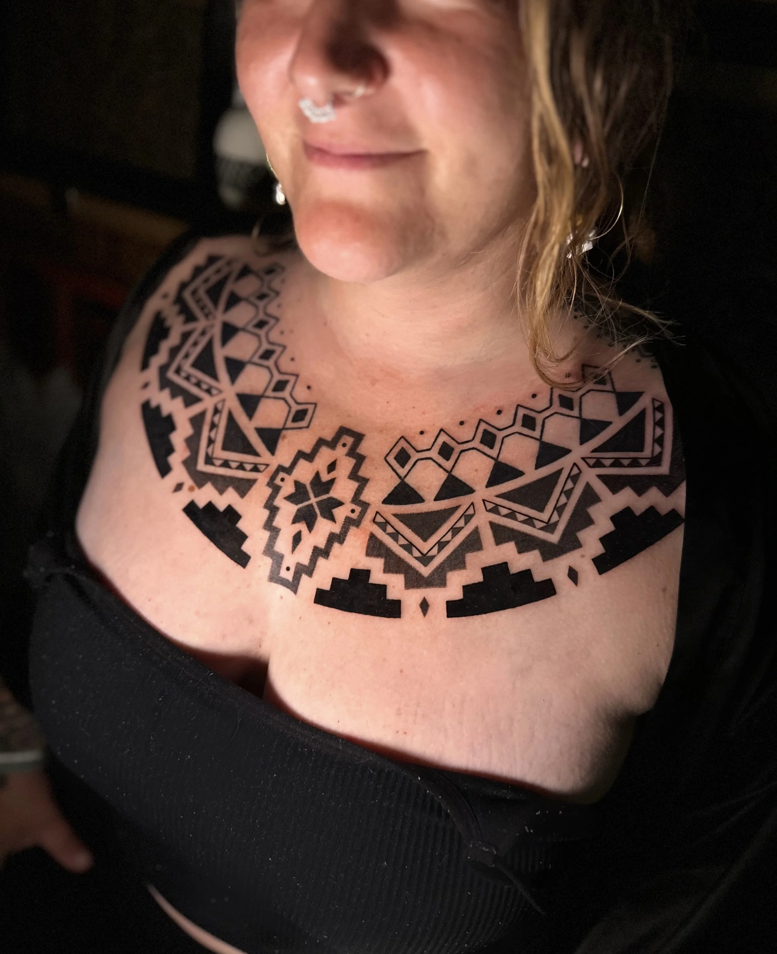 Close-up of a woman's face partially visible, showing her mouth, nose, and curly hair. She has a nose piercing with a silver ring and wears earrings. She has a tattoo with black geometric patterns on her chest and shoulder area, and is wearing a blac