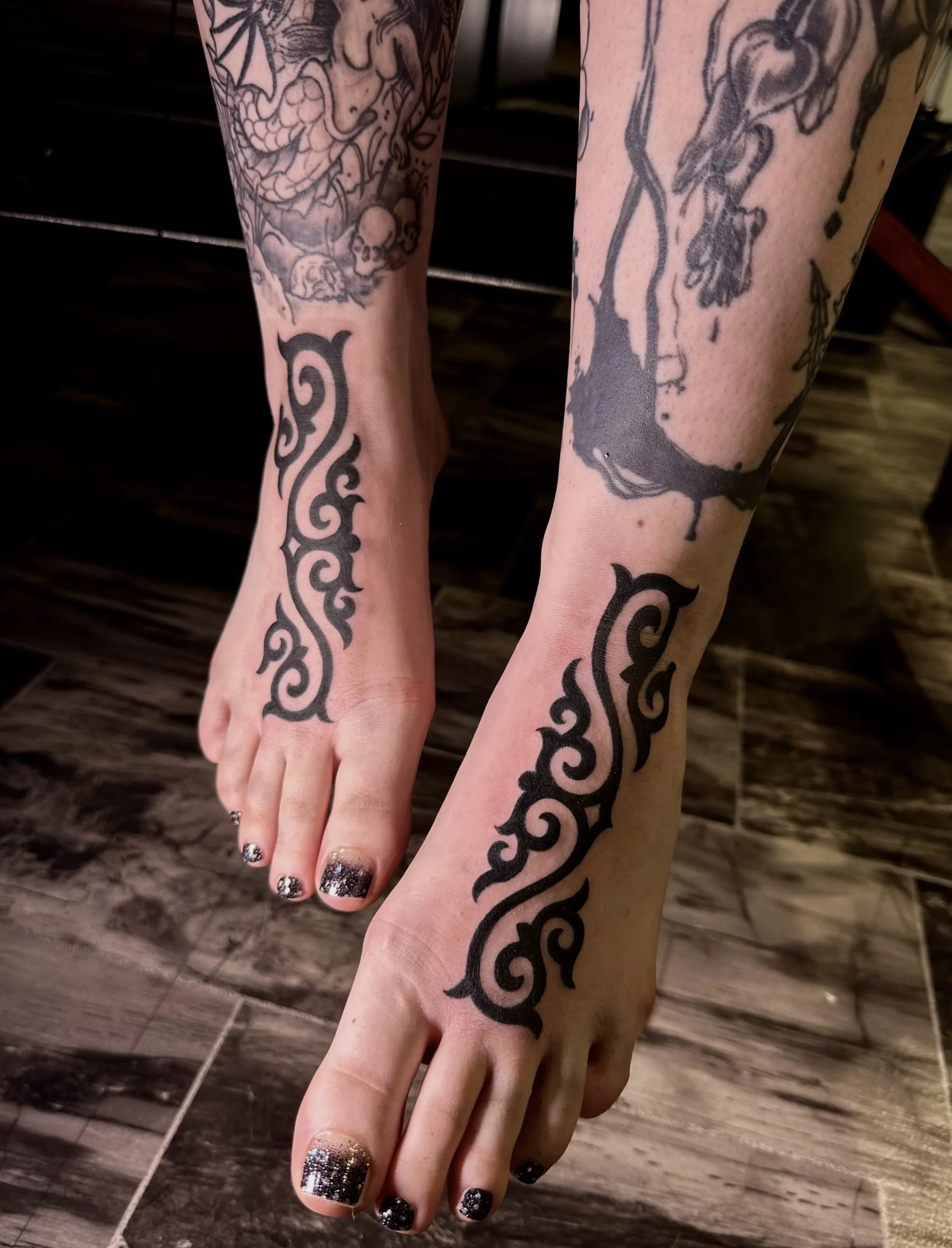 Close-up of two hands with tattoos, one of a stylized black tribal design running along the wrist and forearm, and the other with intricate line drawings including a creature with a skull and other detailed elements, on a wooden floor background.
