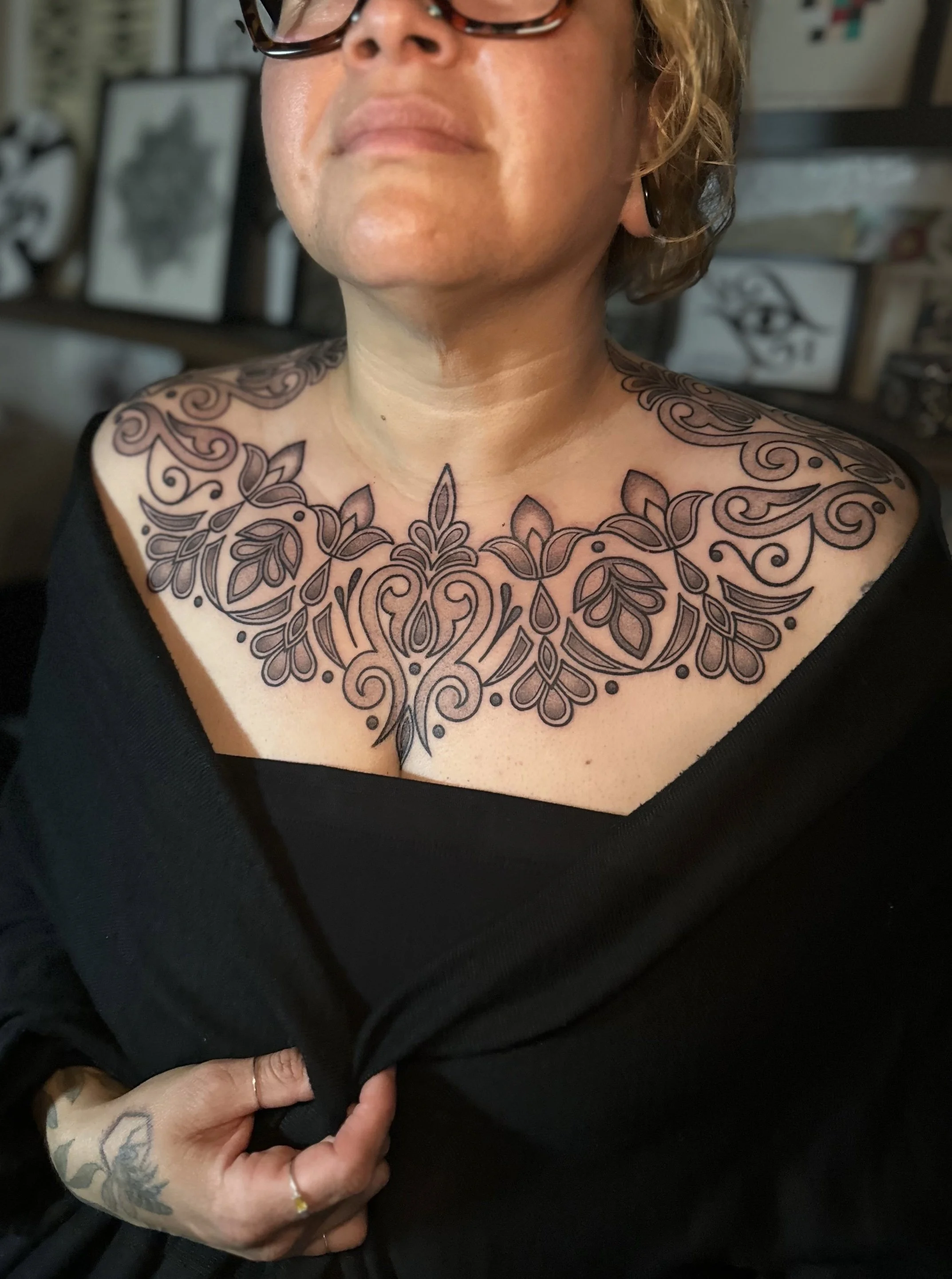 Close-up of a person with glasses who has tattoos on their chest and hand. The person is pulling down their black top to reveal the tattoo. The tattoos are ornate, floral design.