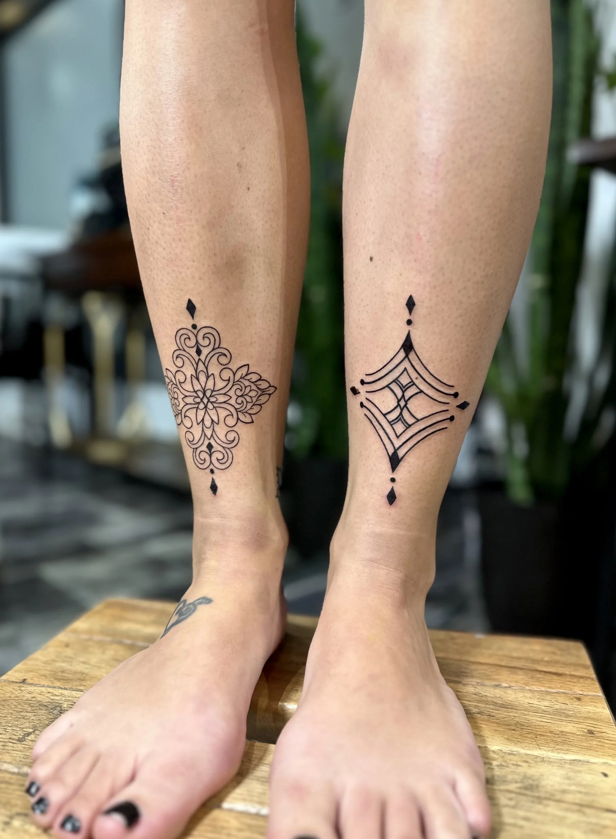 Close-up of two legs with black ink tattoos sitting on a wooden surface, with another person’s bare feet visible in the foreground, and a background with green plants and interior furniture.