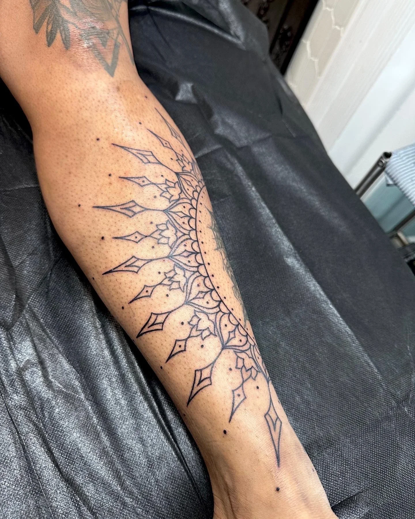 A person's forearm with a detailed black outlines tattoo of a geometric floral mandala design, lying on black leather surface.