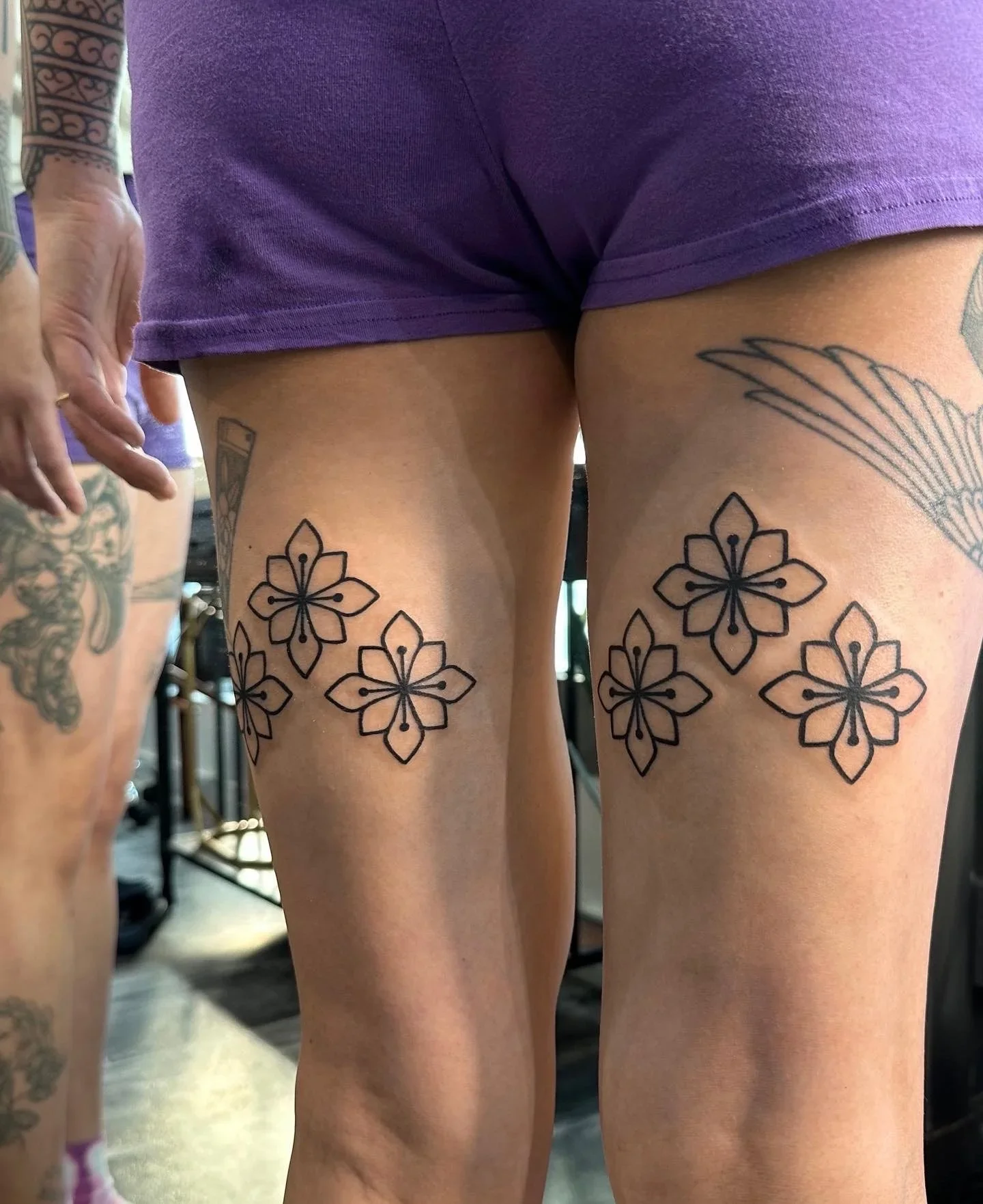 Close-up photo of two women with tattoos on their thighs, wearing purple shorts, in a gym setting.