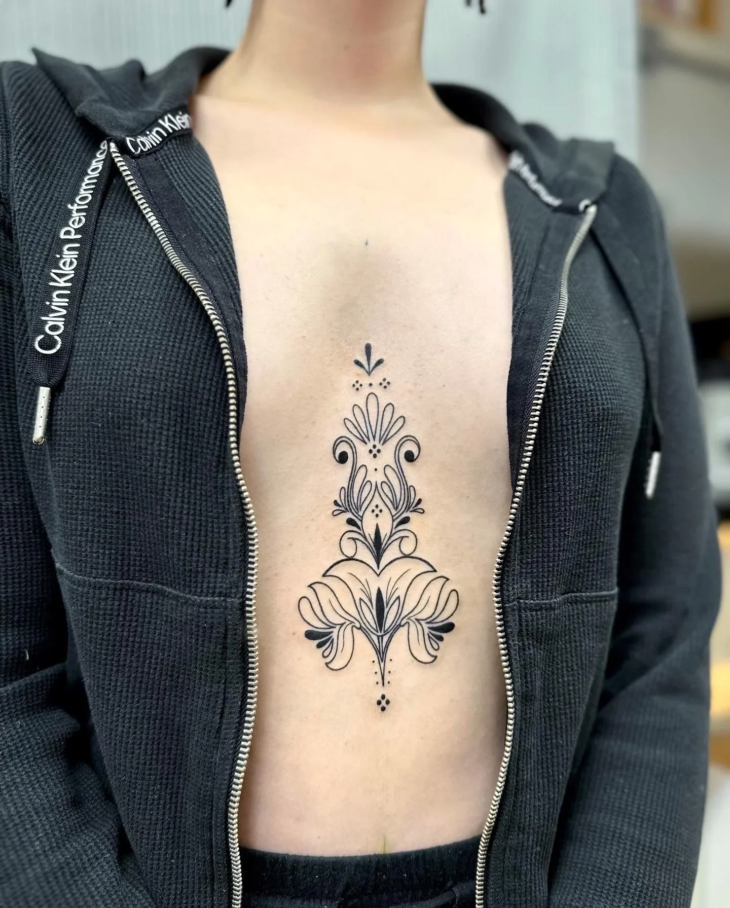A tattoo with black ink adorns the center of a person's chest, featuring symmetrical, ornamental floral and geometric patterns.