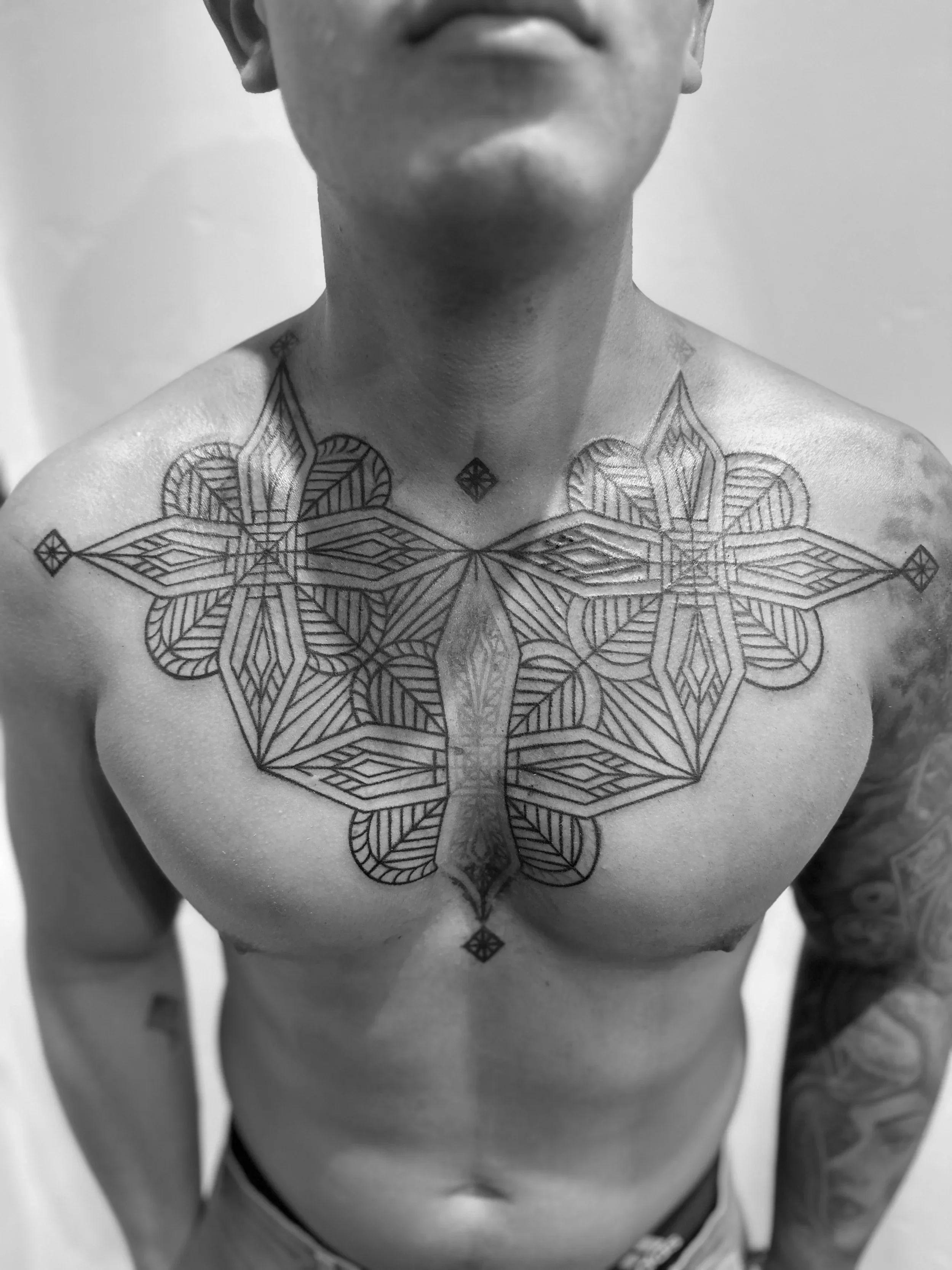 Black and white photo of a man with a geometric chest tattoo, mostly showing his torso and part of his face, with a serious expression.