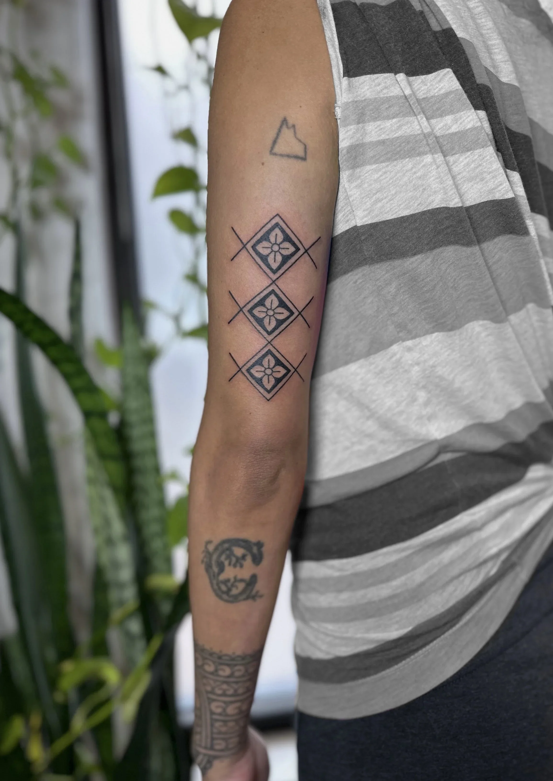 A person's upper arm with three tattoos: an outline of a fox head, and a design of three diamond-shaped patterns with floral motifs inside, arranged vertically. The person is wearing a gray and black striped sleeveless shirt.