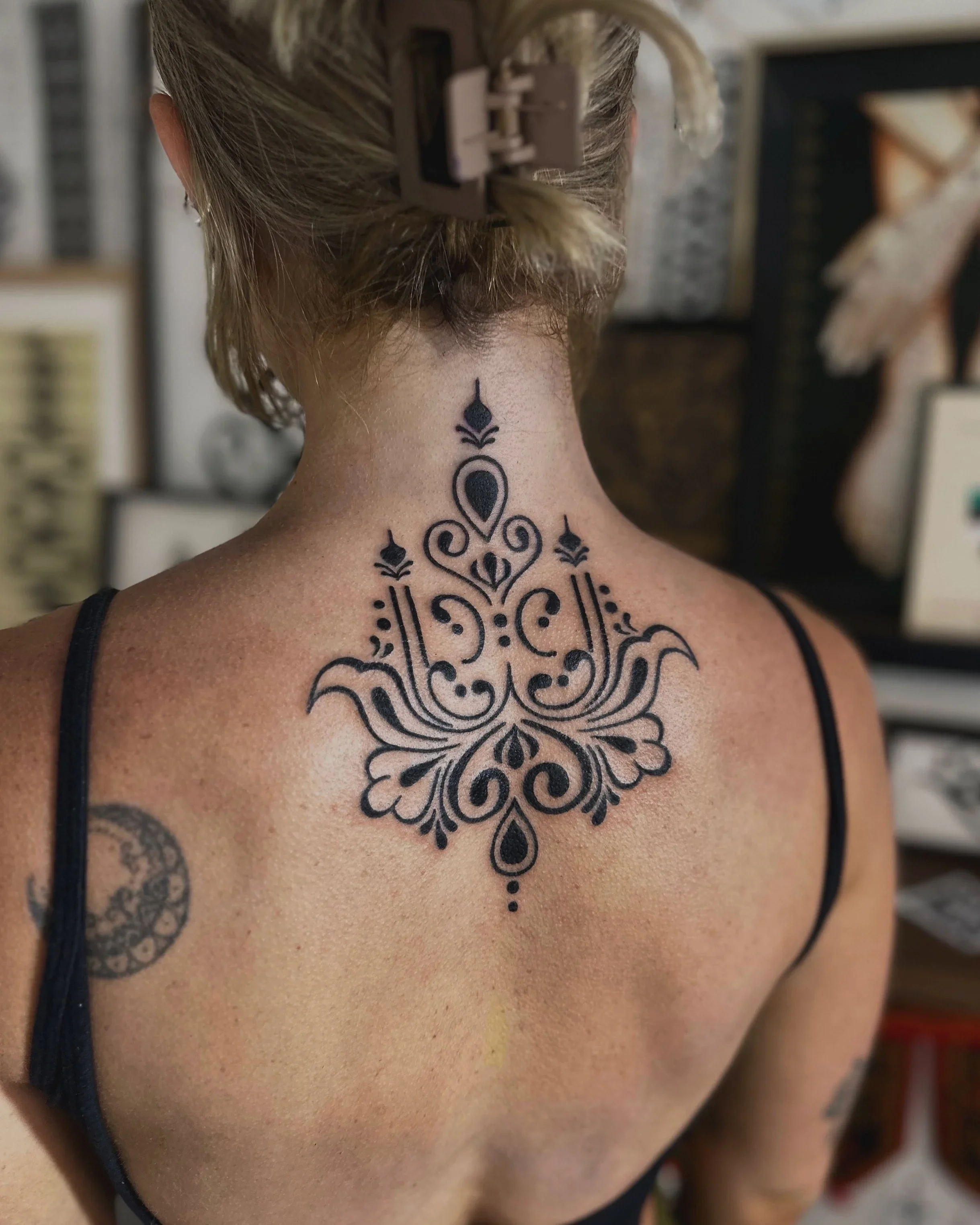 Back of a person with light skin, wearing a black strap top, revealing a large intricate black tattoo on the upper back and neck, with hair pulled up using a beige hair clip, set in a blurred indoor background.