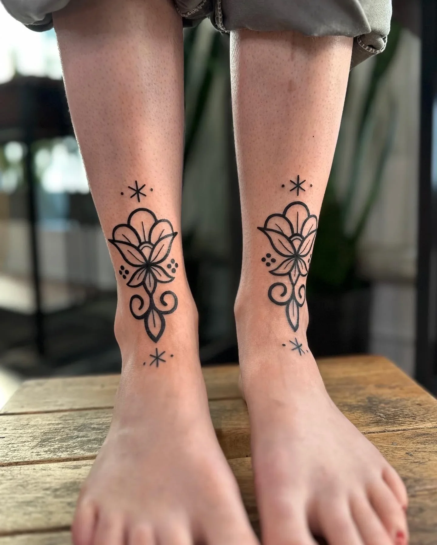 Pair of feet with matching black line tattoos of a lotus flower on the inner ankles, with decorative dots and stars, on a wooden surface.