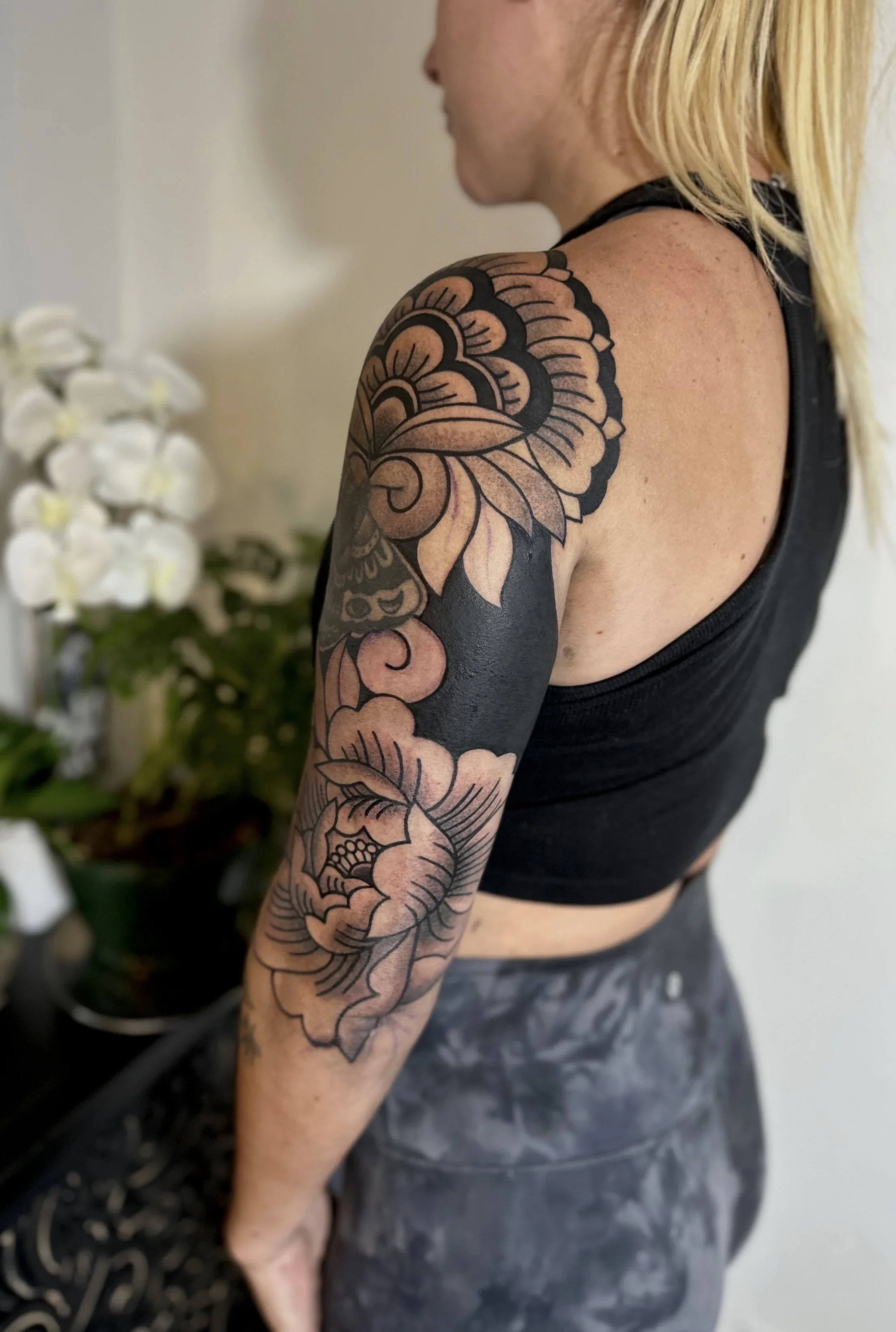 A person with a large floral tattoo on their upper arm and shoulder, wearing a black sleeveless top and gray patterned shorts, standing indoors near a white wall.