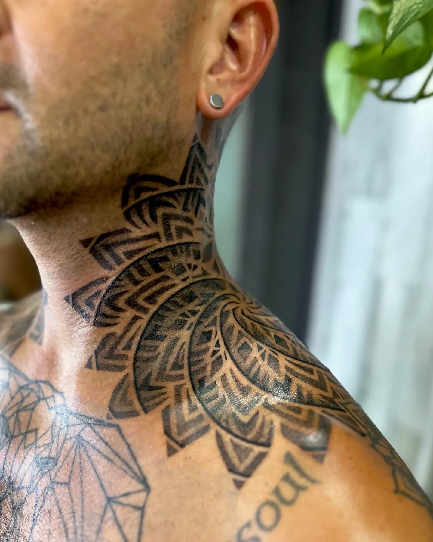 Close-up of a person's shoulder and neck featuring extensive black ink tattoos with geometric patterns and text, alongside a silver stud earring in the left ear.