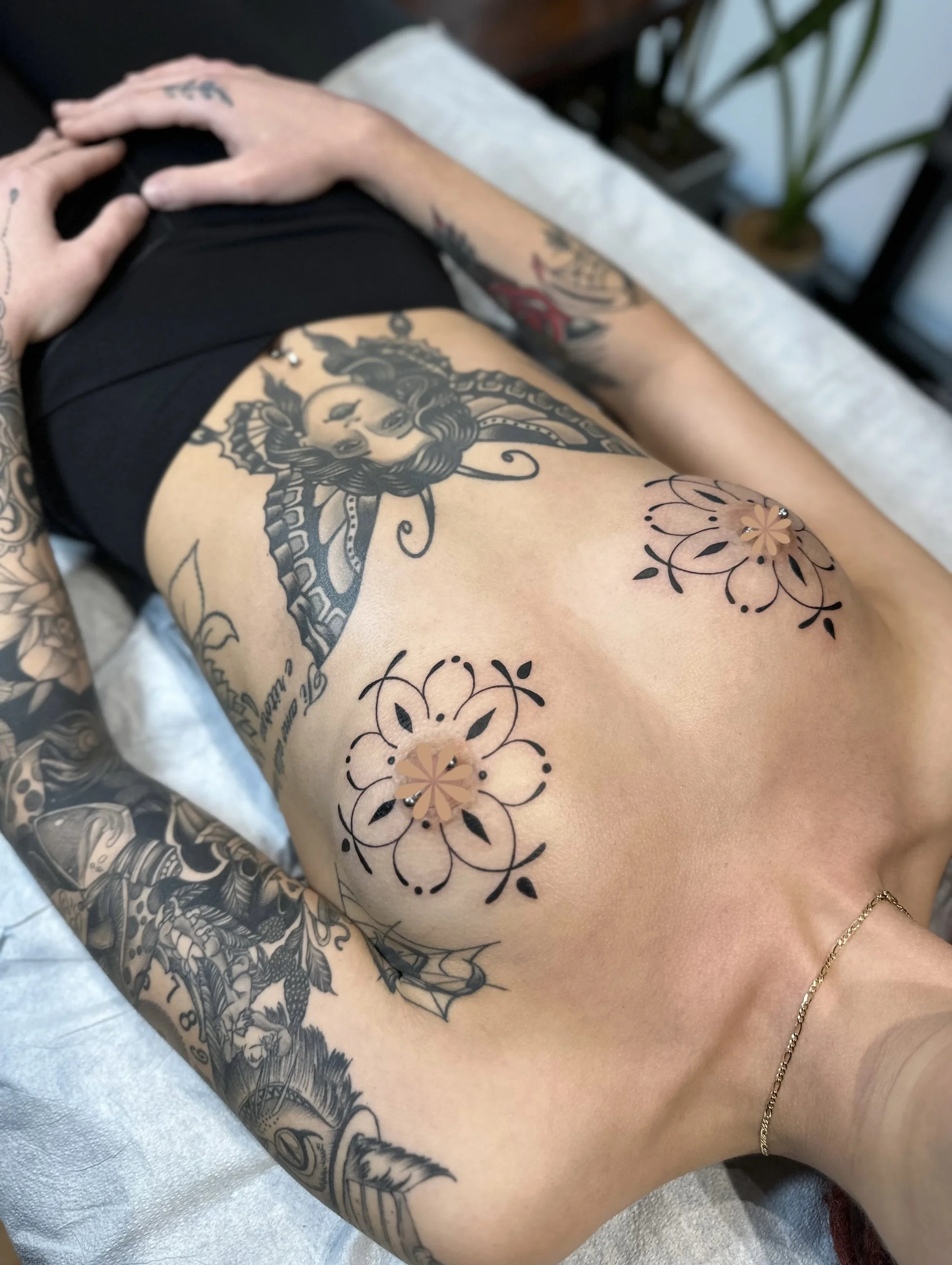 A person's arm with various tattoos, including a large portrait of a woman with curly hair and facial features, a floral design, and other decorative elements. The person is lying on an examination table with a white paper sheet, wearing a black shir
