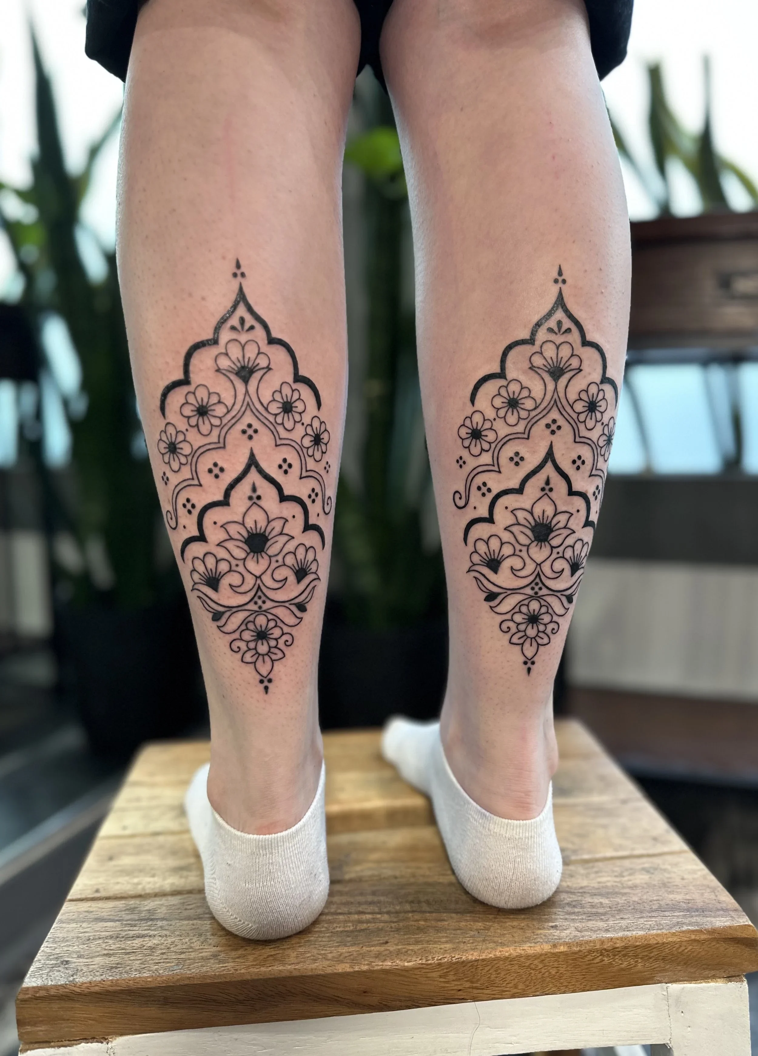 A pair of mannequin legs with black tattoo designs of floral patterns on the calves, standing on a wooden surface with green plants and furniture in the background.