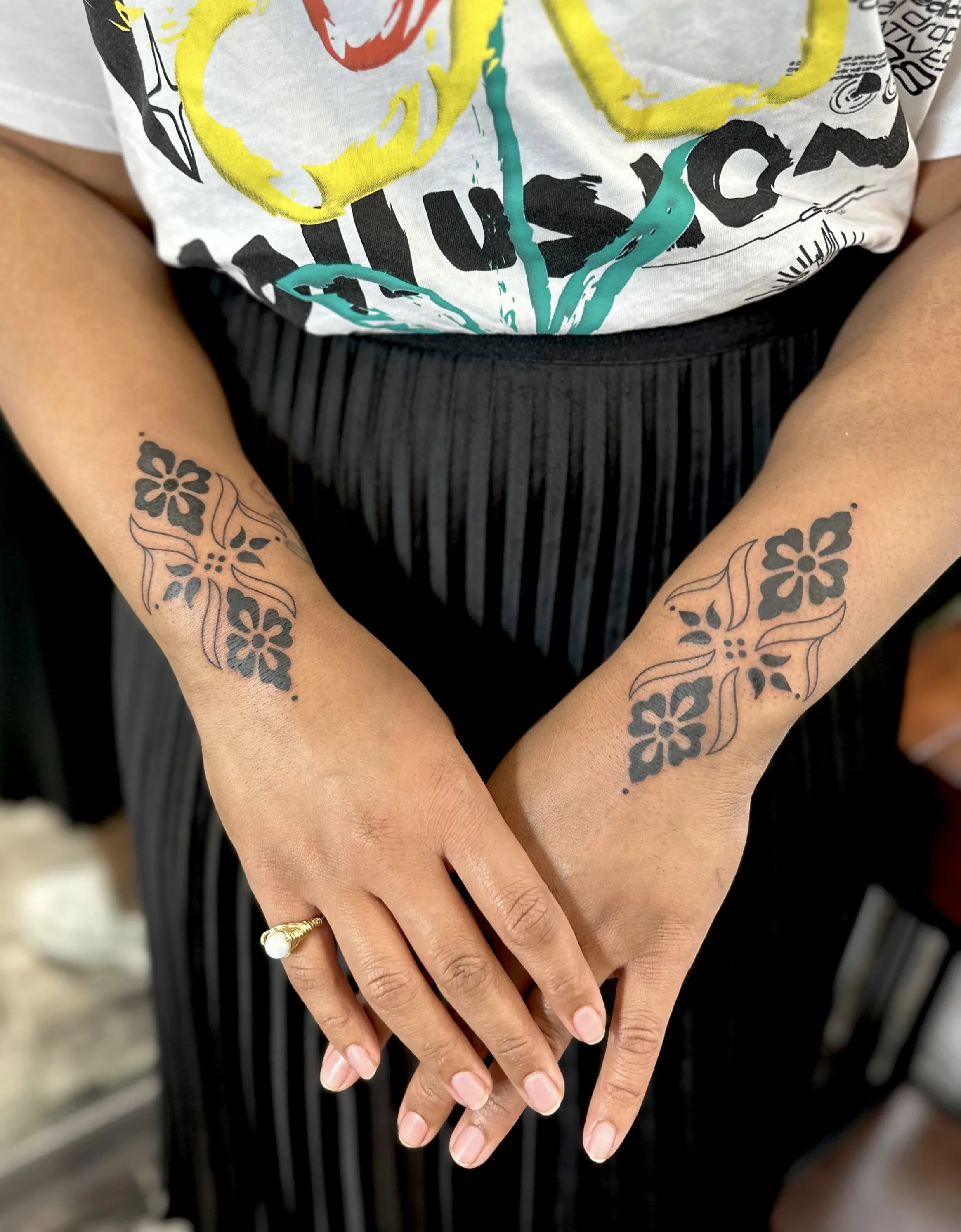 Close-up of two hands with floral tattoos, intertwined, with a gold ring on the ring finger of the left hand, against a background of black and white striped pants and a white shirt with colorful abstract designs.