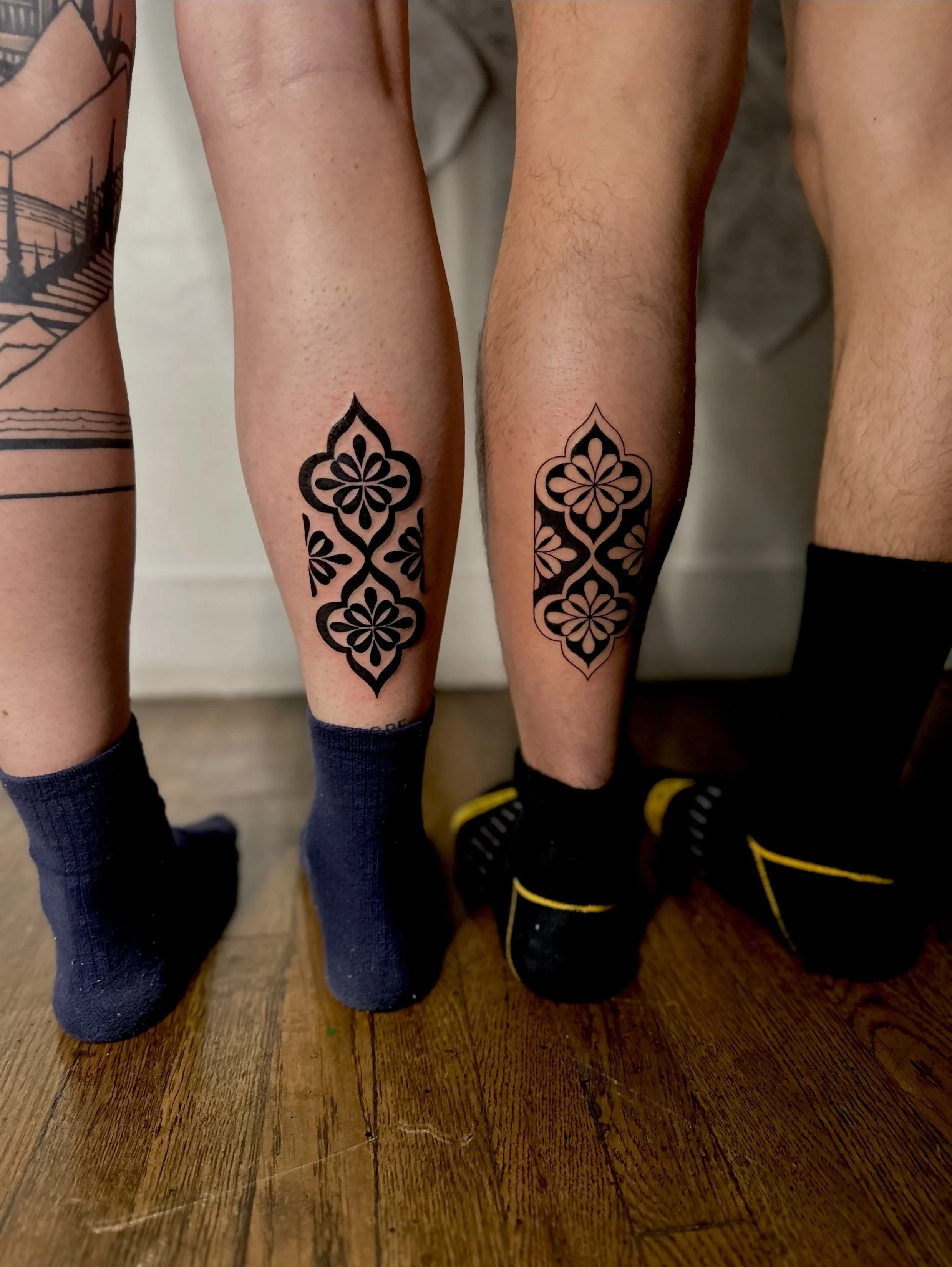 Close-up of four people's legs standing on a wooden floor, showing their socks and tattoos on their legs. Two people have matching black tattooed crowns on their calves.