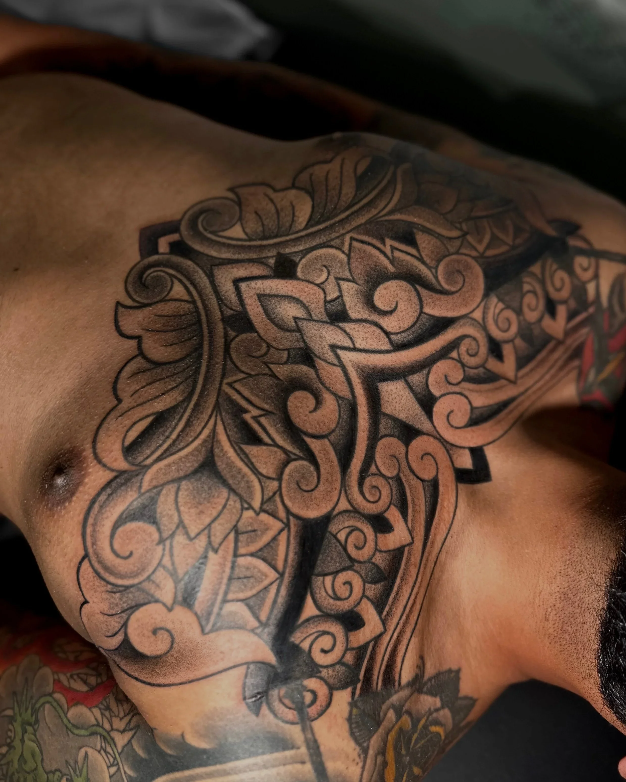 A close-up of a person's shoulder and chest showing an intricate, black-and-gray tattoo with floral and abstract patterns.