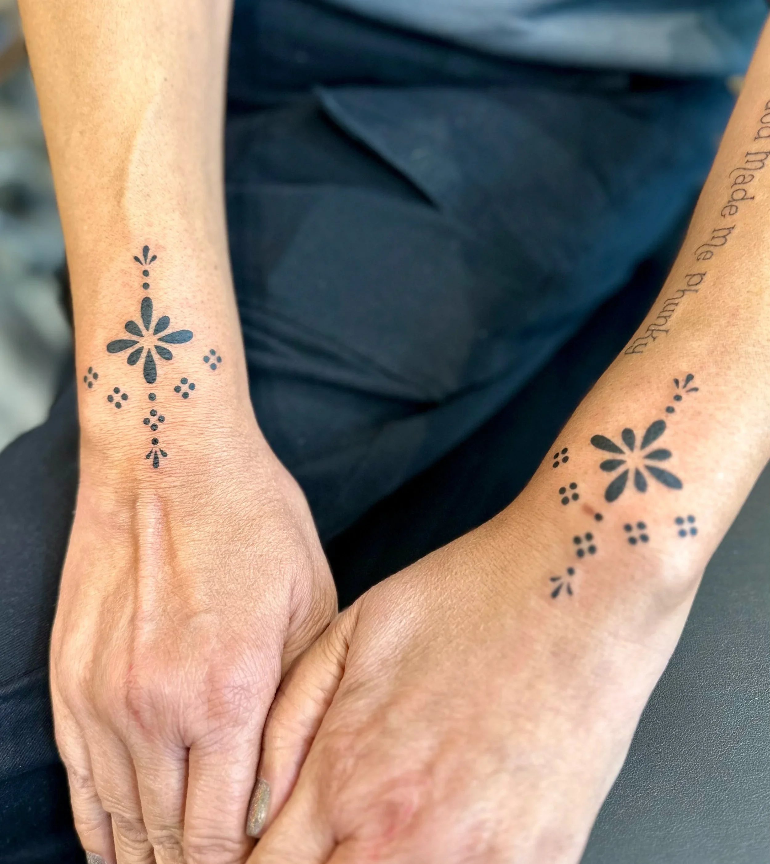 Close-up of two hands pressed together, showing tattoos and jewelry on a person's arm and hand, with a dark background.
