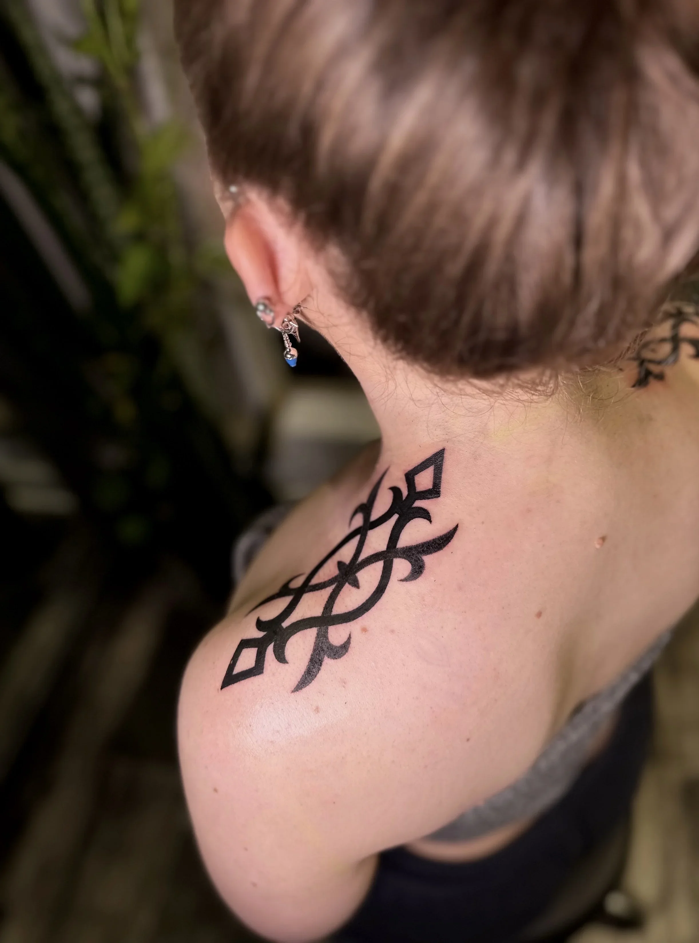 A close-up of a person's shoulder, showing a large black tattoo and a silver earring with a blue pendant. The person has brown hair and is wearing a gray top. The background is blurred with green and darker colors.