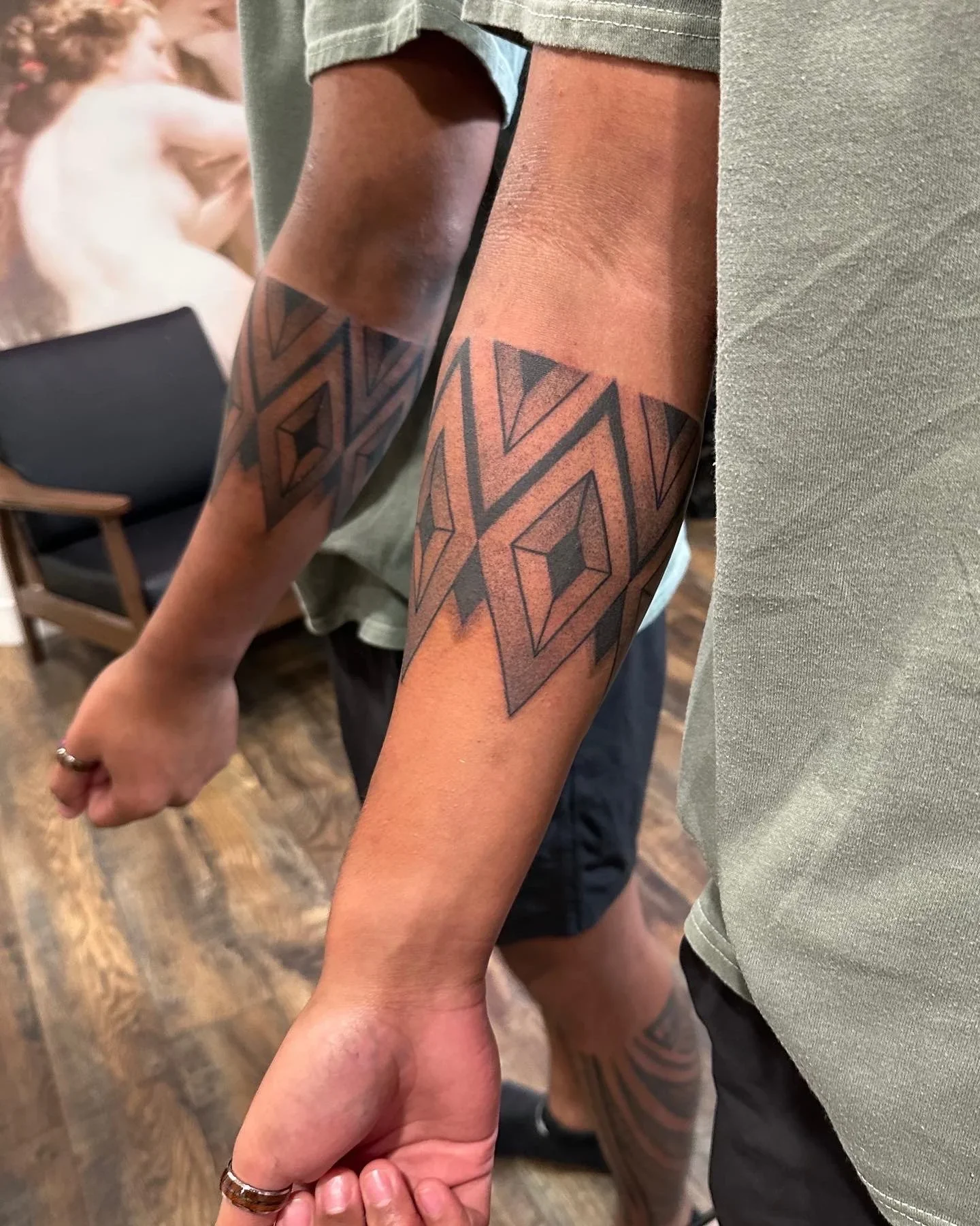 Two people holding hands, showing tattoos on their arms. One person's arm has a geometric tattoo with black ink, and the other's arm has a similar geometric tattoo. The setting appears to be indoors with a wooden floor and a chair in the background.