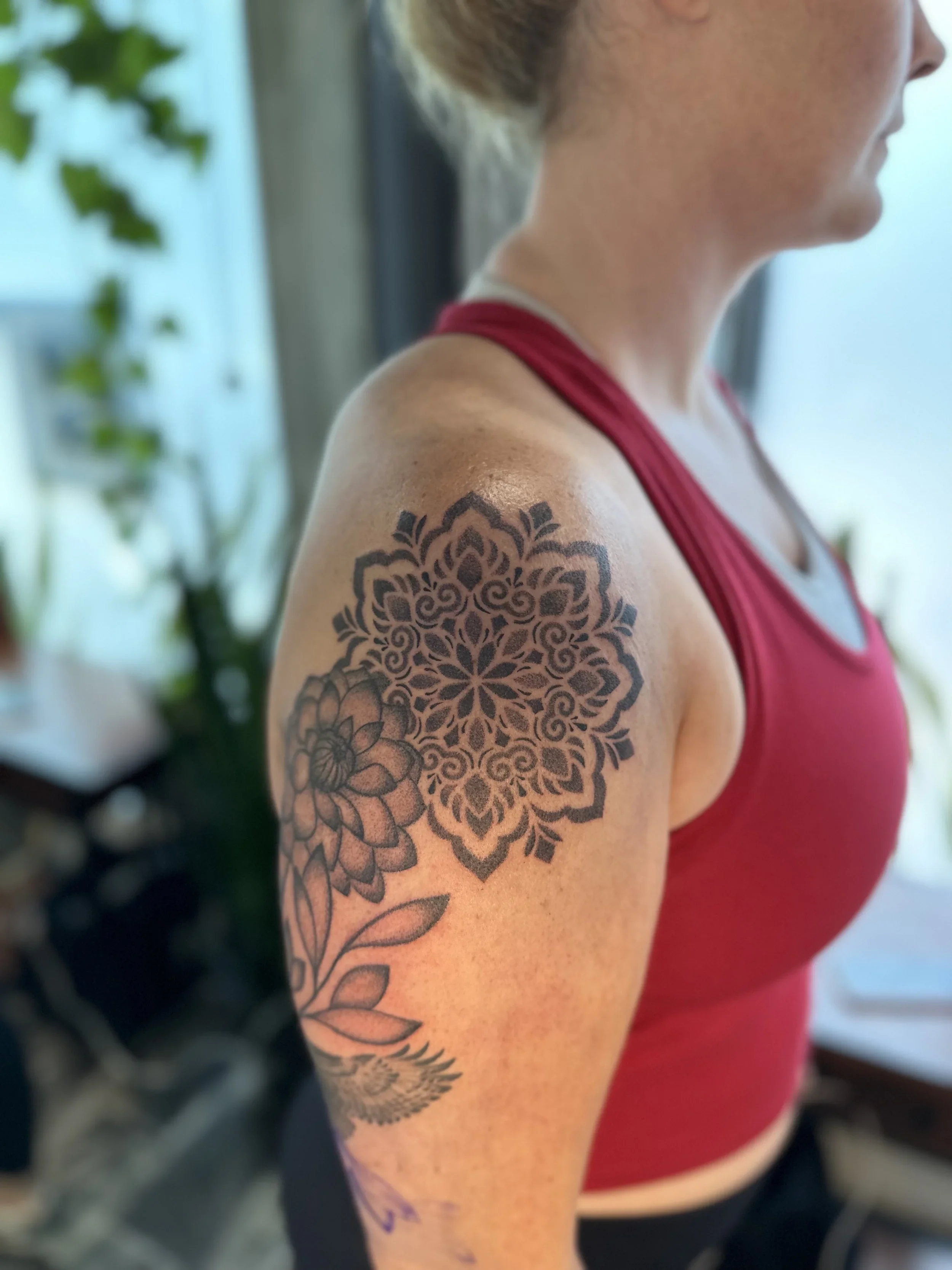 Close-up of a woman wearing a red tank top, showing a detailed black floral tattoo on her upper arm, with green plants in the background.
