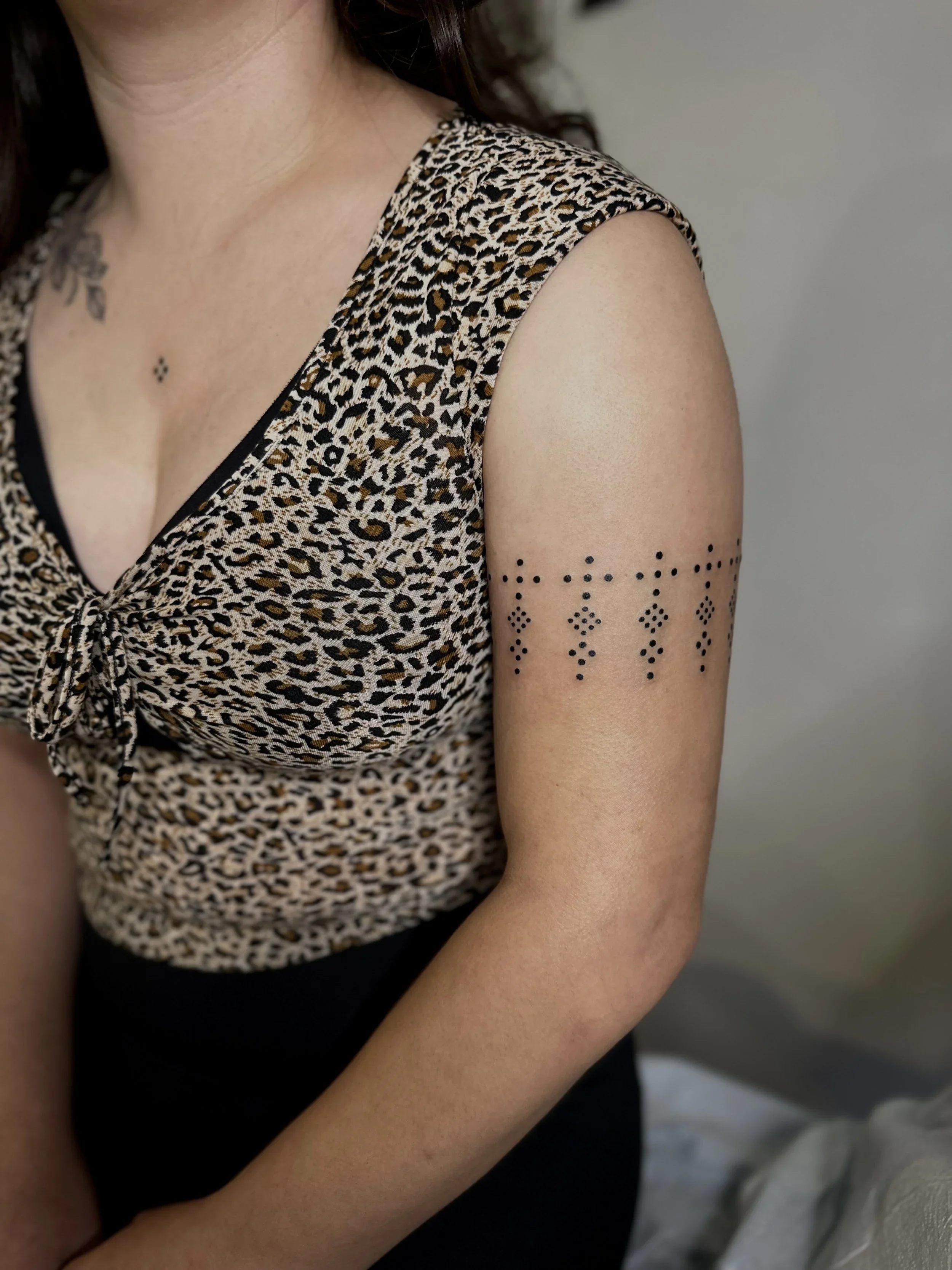 Close-up of a woman's shoulder and upper arm with small black diamond-shaped tattoos, wearing a sleeveless leopard print top.