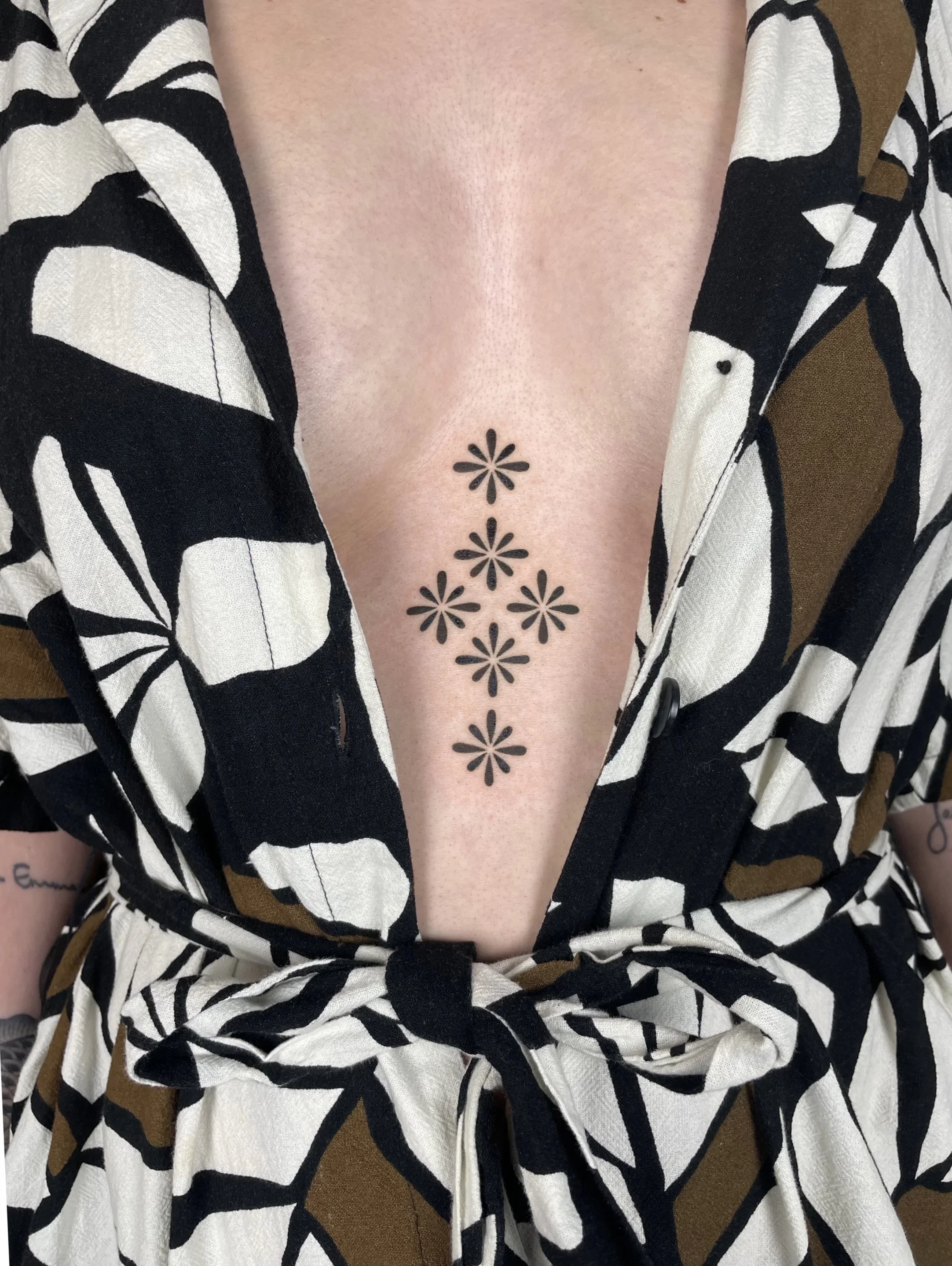 Close-up of a woman's chest with a black floral tattoo between her breasts, visible above the open collar of a black and white patterned shirt.