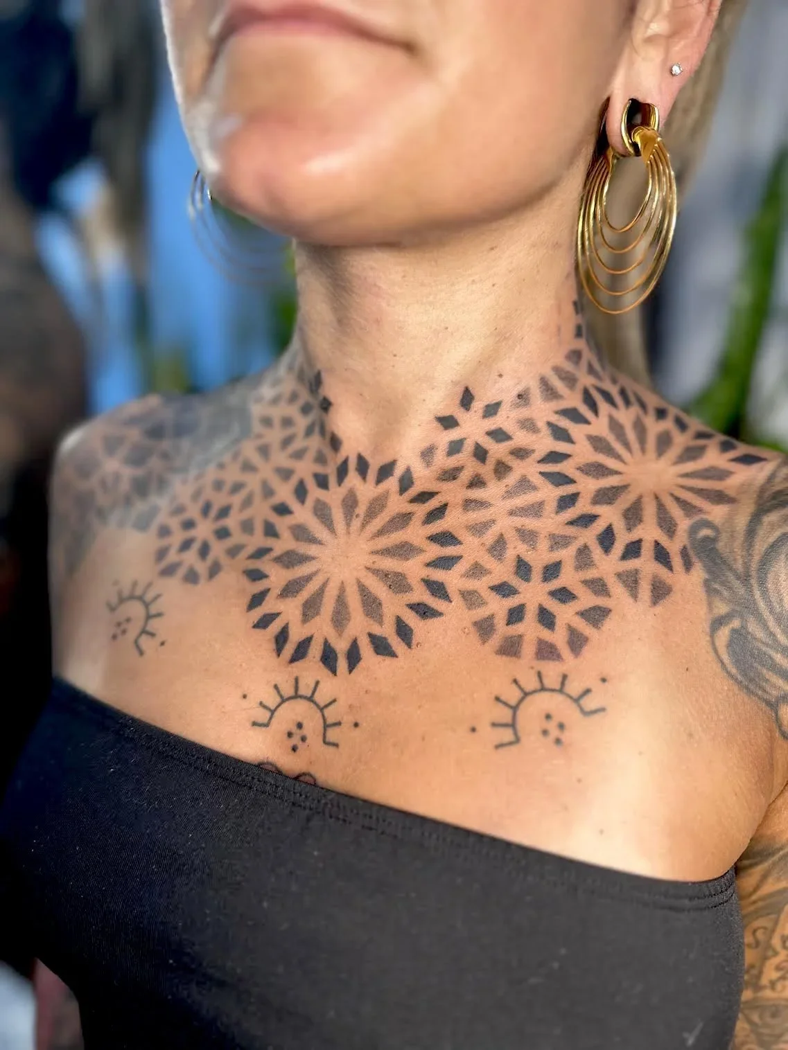 Close-up of a woman's chest and neck with large geometric tattoo patterns and earrings.