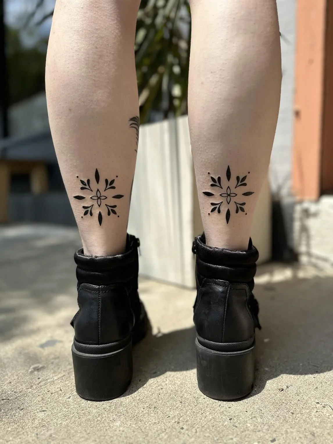 Close-up of a person's legs with black floral tattoos on the lower calves, wearing black platform boots, outdoors on a sunny day.