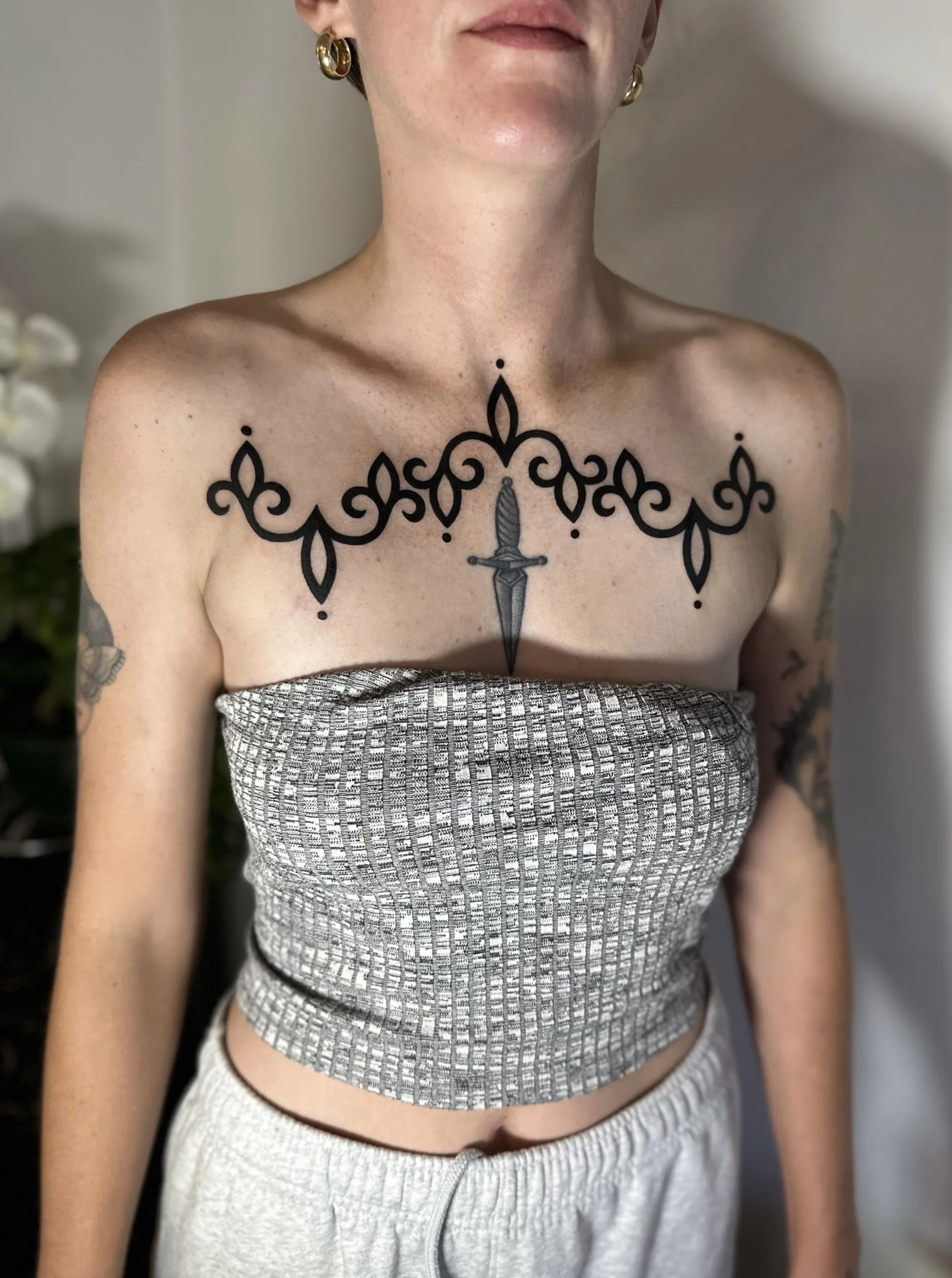 A woman with tattoos on her arm and chest wearing gold hoop earrings and a strapless patterned top, standing indoors.