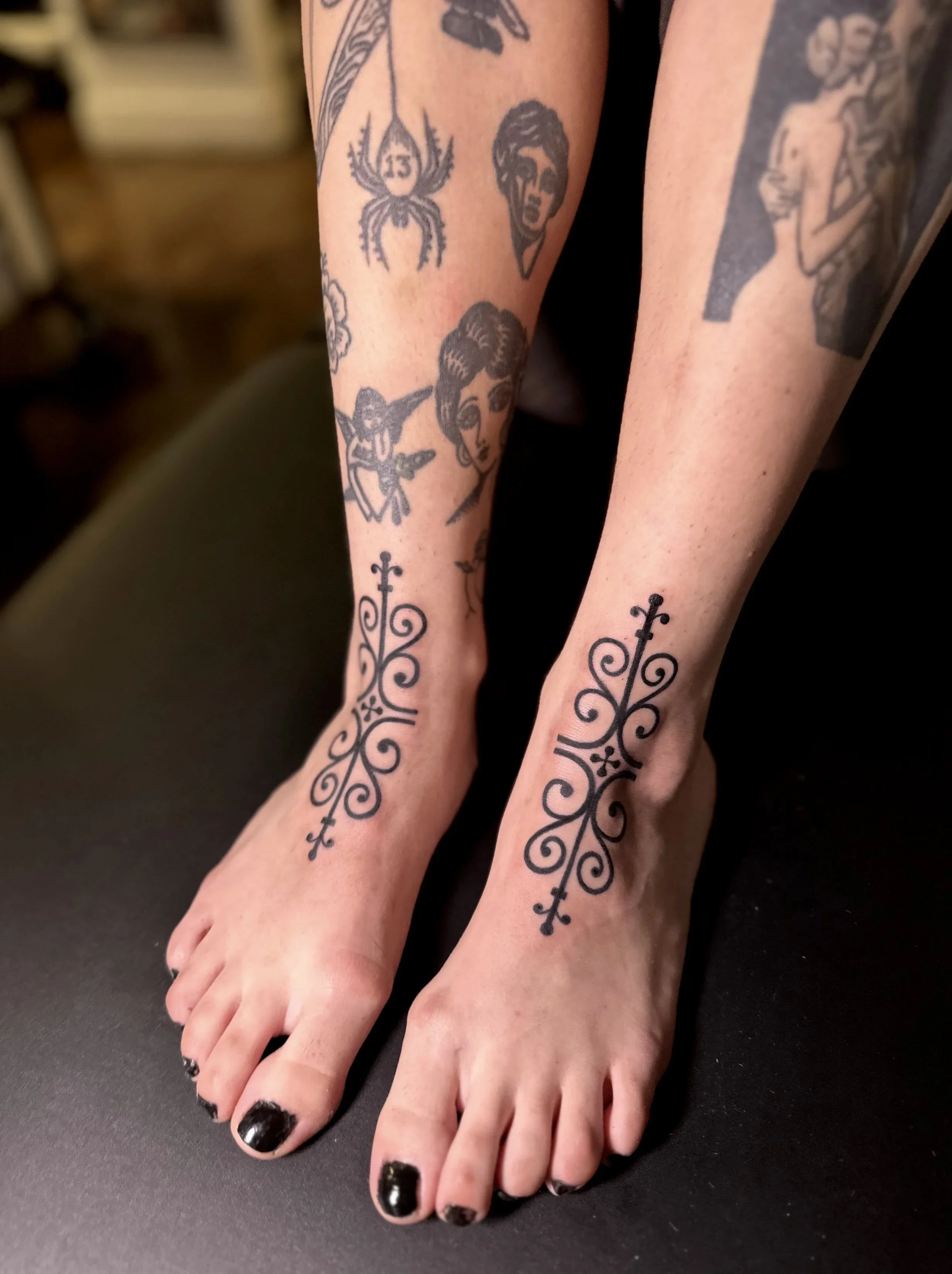 A person's lower legs and feet with multiple tattoos, including a woman's face, a spider, a wolf head, and various decorative designs. The person has black nail polish.