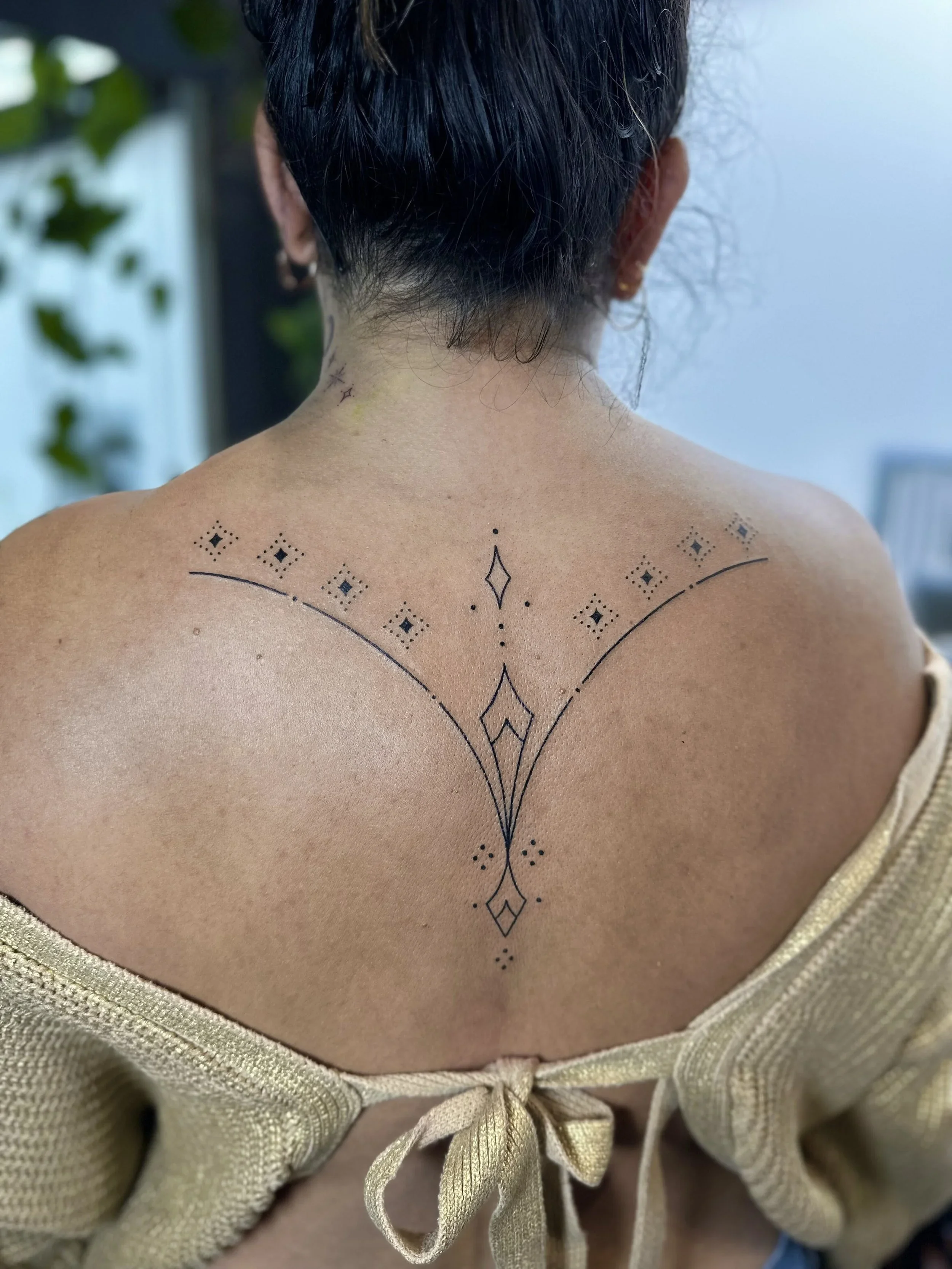 Back view of a woman with a tattoo on her upper back and neck, wearing a beige top with a bow at the back.