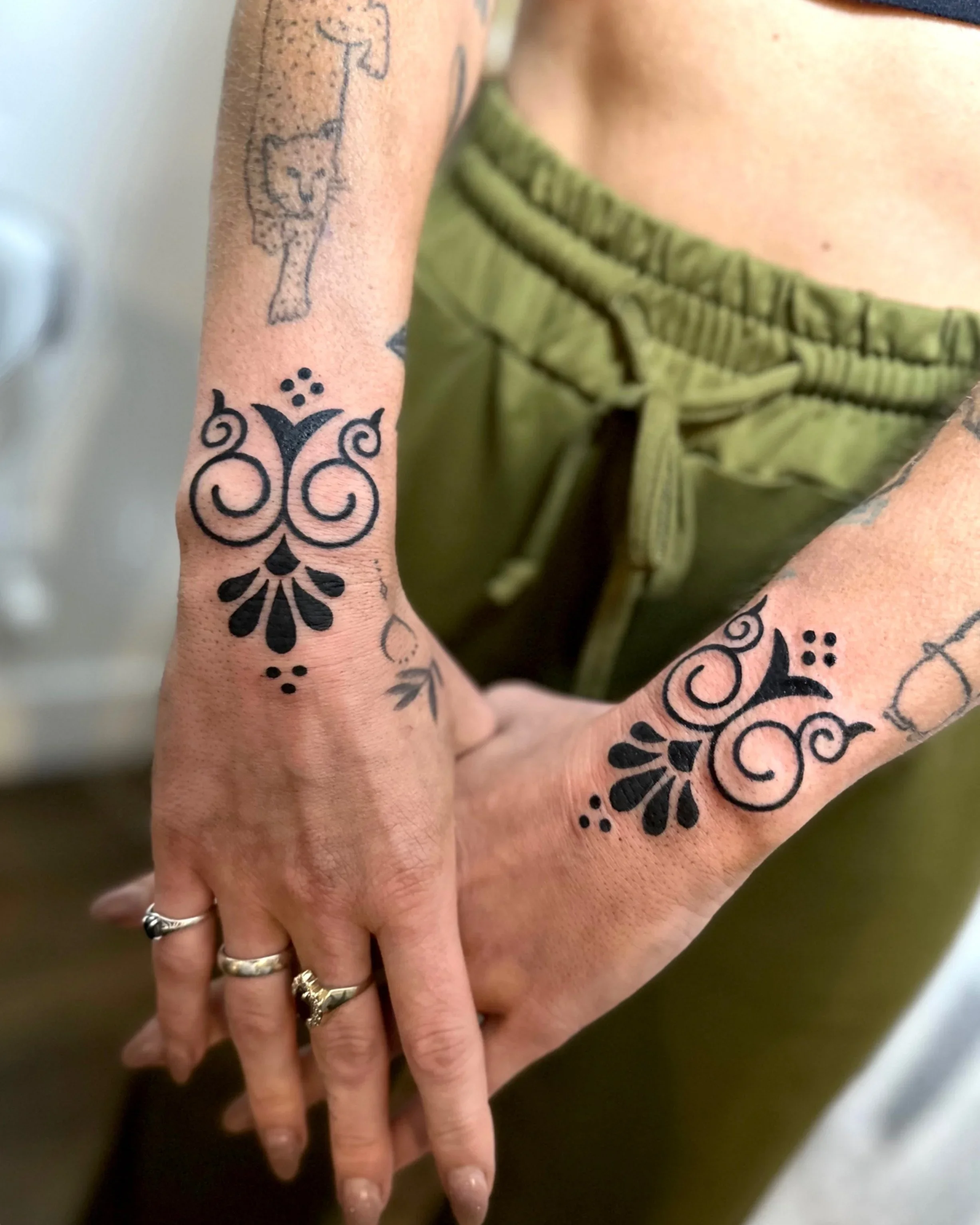 Close-up of a person's hand with tattoos and silver rings, fingers intertwined over a surface, wearing olive green pants with a drawstring.