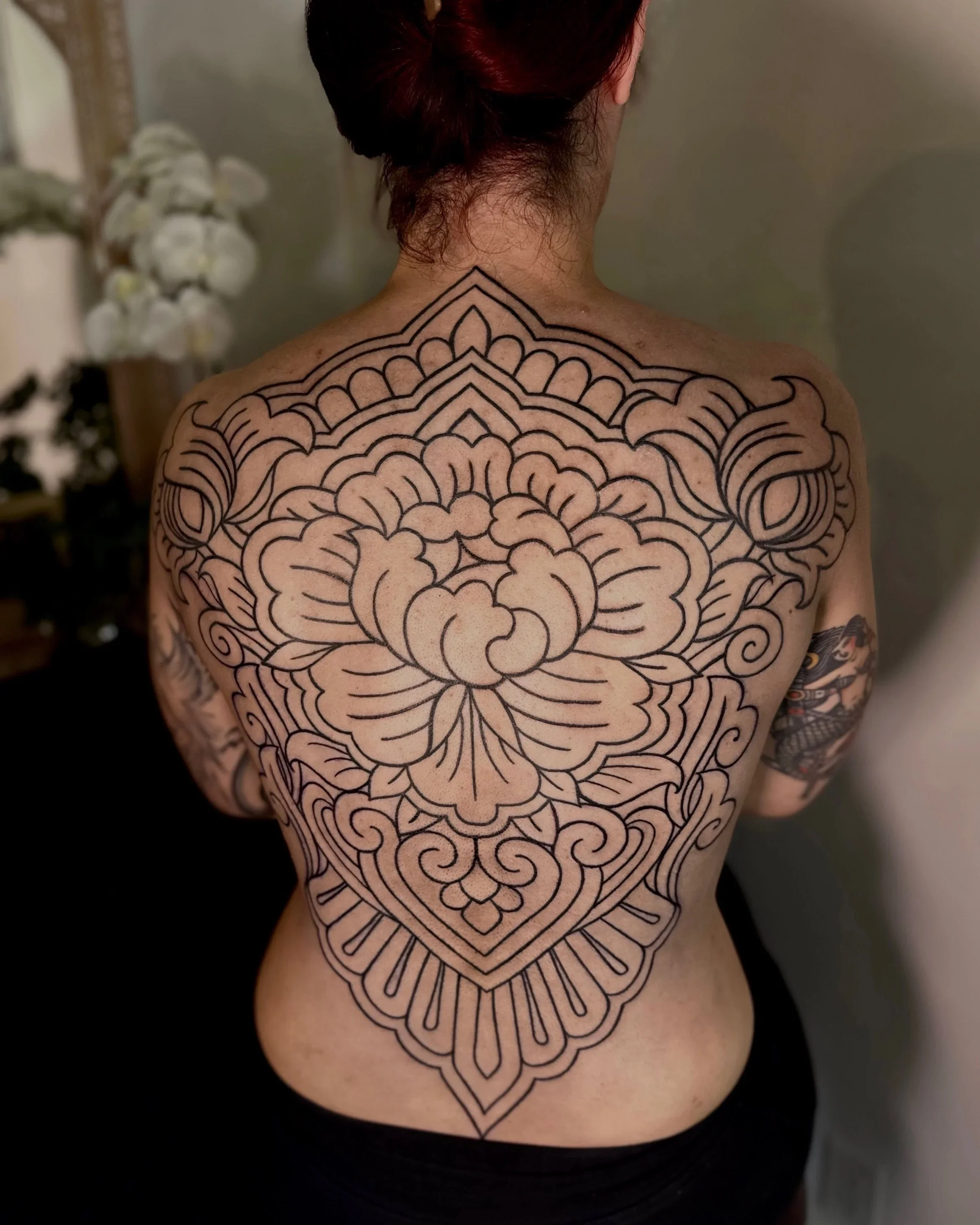 A person with red hair styled in a bun, showing their back and shoulders, with a large, intricate black line tattoo of a mandala and floral design covering the upper back, shoulders, and down the center of the back.