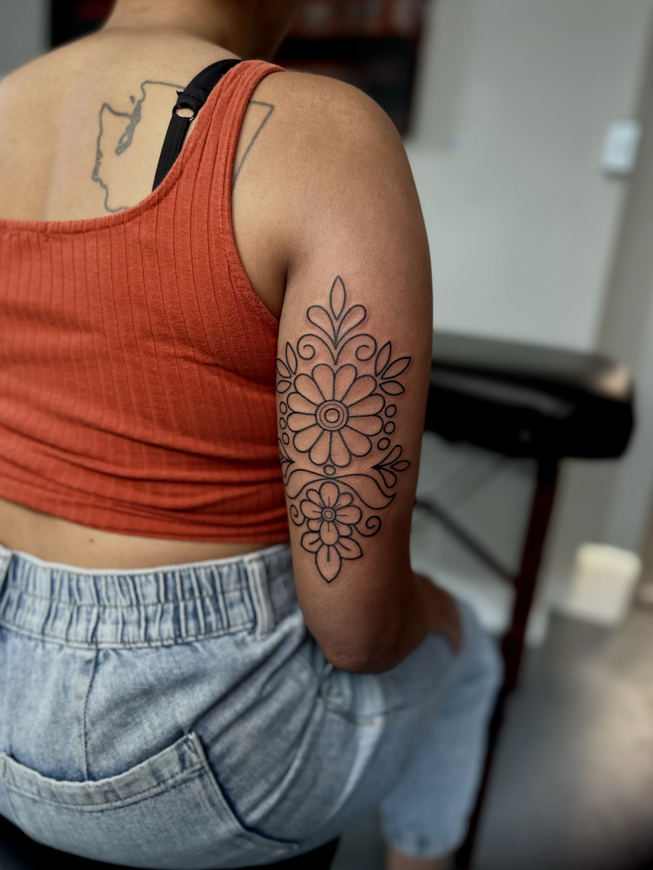 Close-up of a person's upper arm and shoulder, showing a floral tattoo and a map tattoo on the back, wearing an orange tank top and blue jeans.