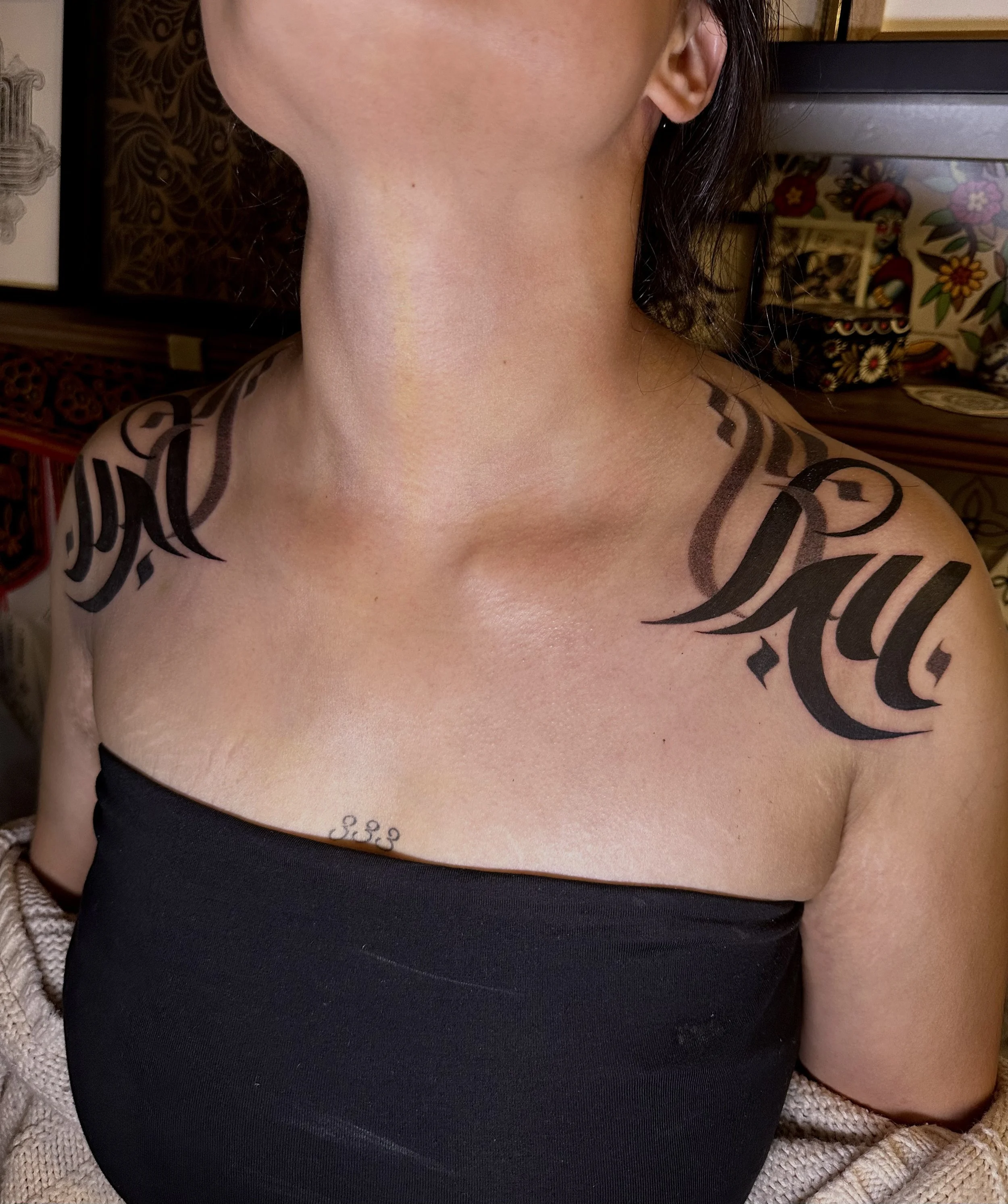 Close-up of a woman's neck, shoulder, and part of her face, showing tattoos on her shoulder and collarbone, wearing a black top with a patterned background.