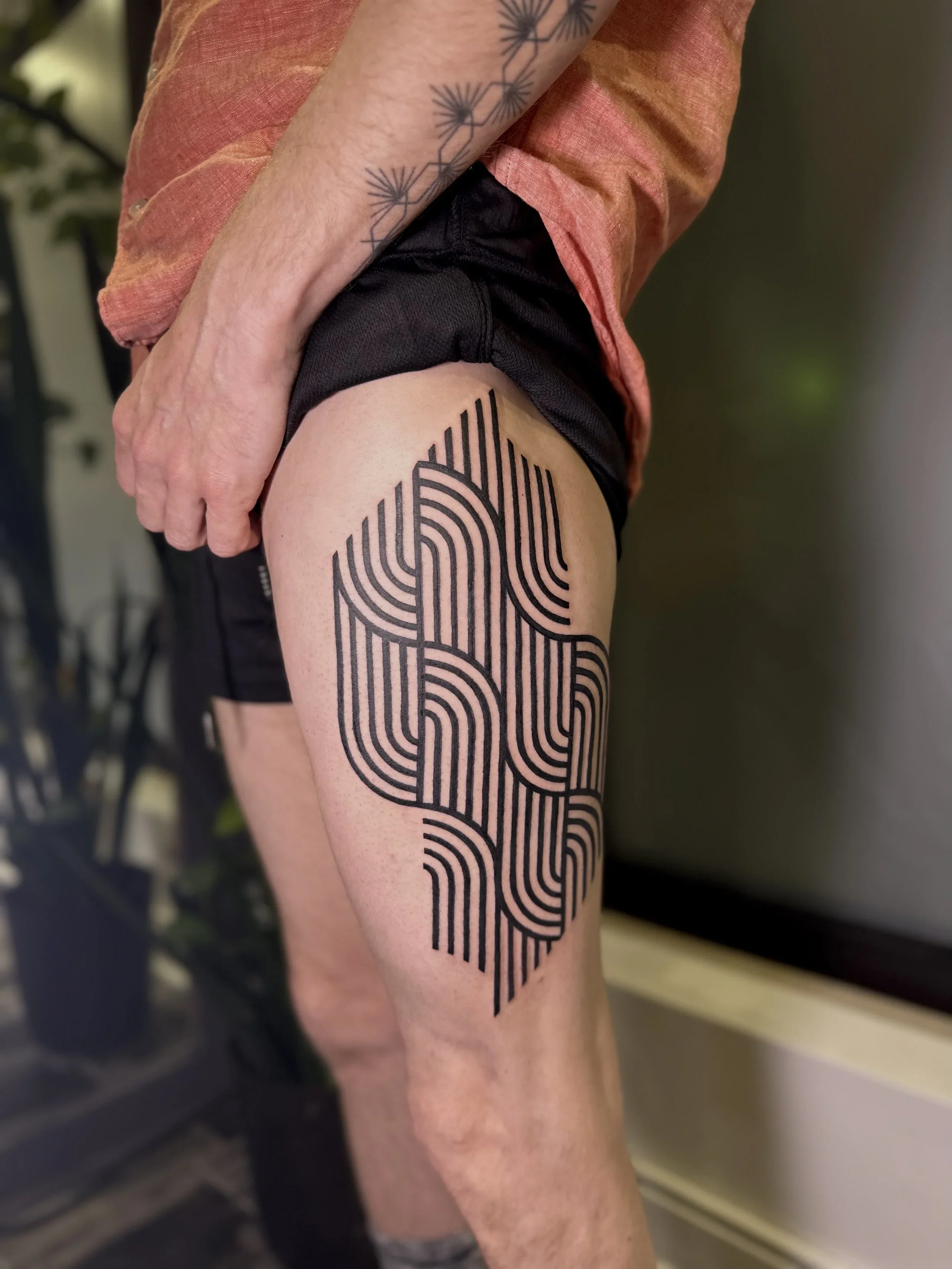 A person's upper leg with a large black tattoo resembling a fingerprint on the thigh, wearing black shorts and a worn pinkish-orange shirt.