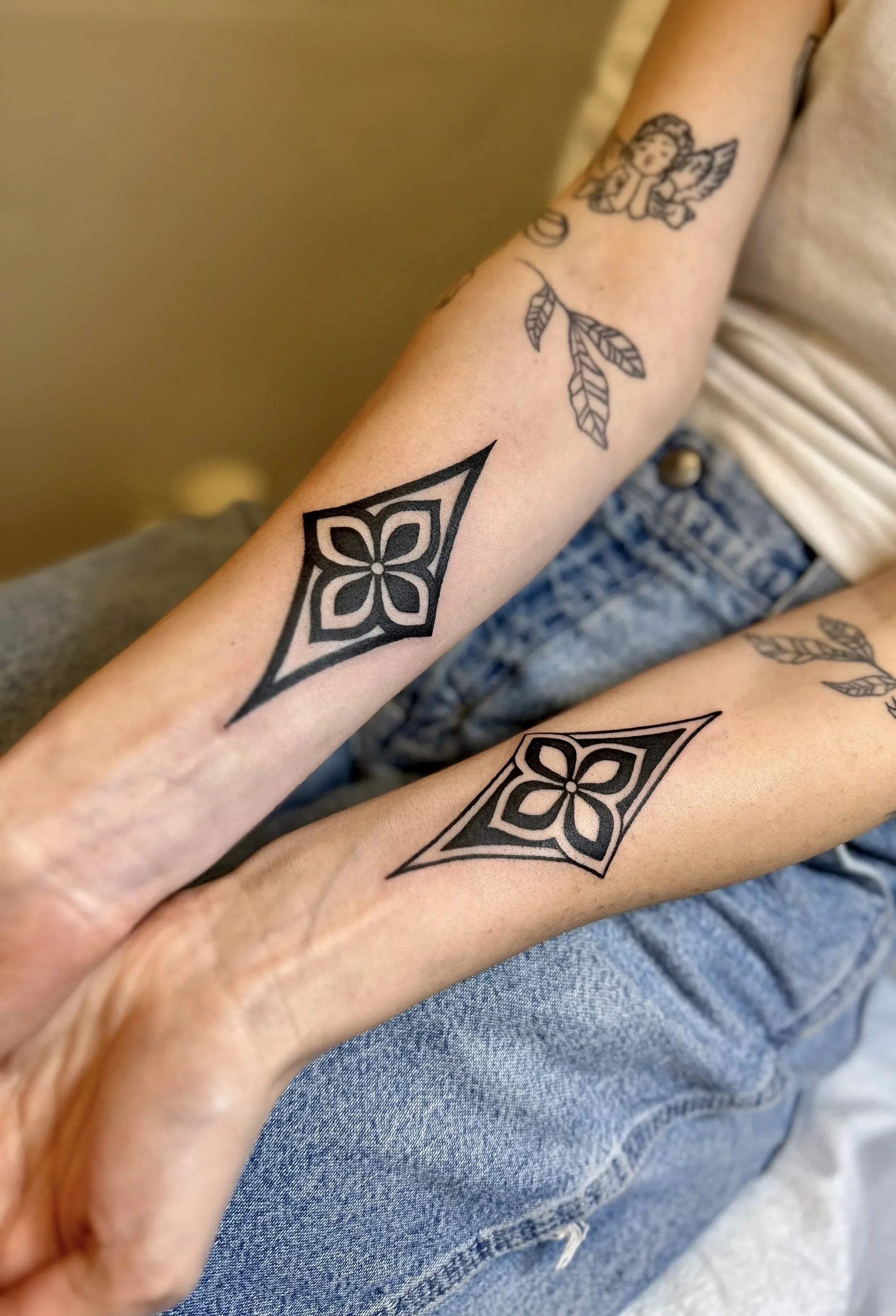 Close-up of a person's arm and hand with multiple tattoos, including a geometric flower design and an angel with wings and a leaf on the arm, with blue jeans and a beige shirt.