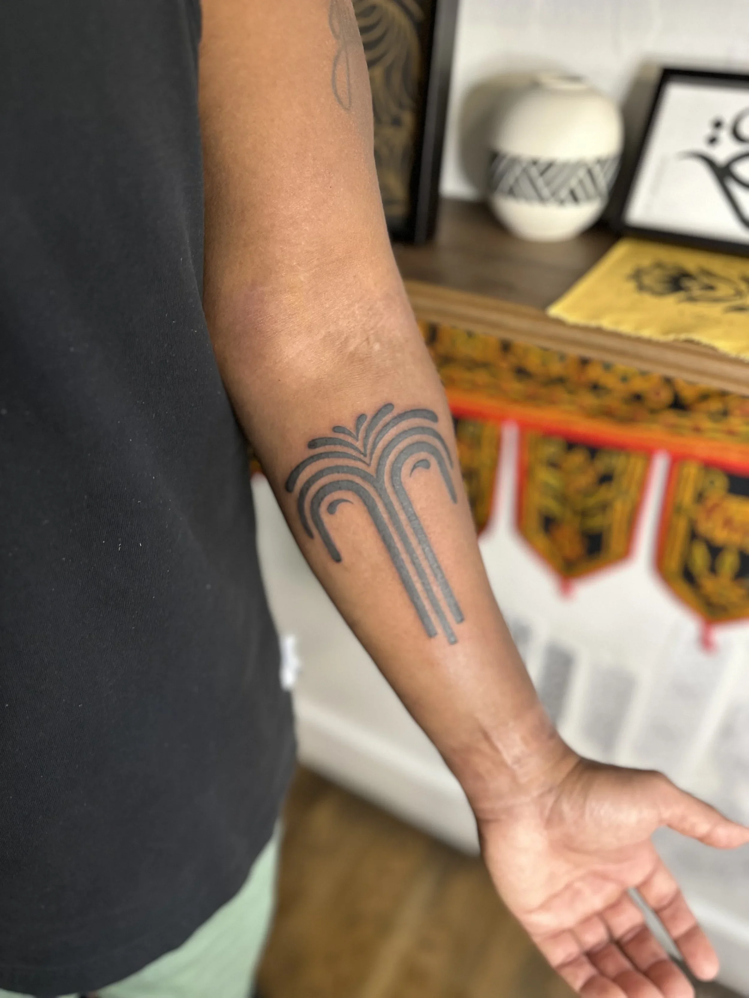 A person's arm with a geometric tattoo, showing the forearm and part of the hand, with a background of framed art and a decorative vessel, upon a patterned table or surface.