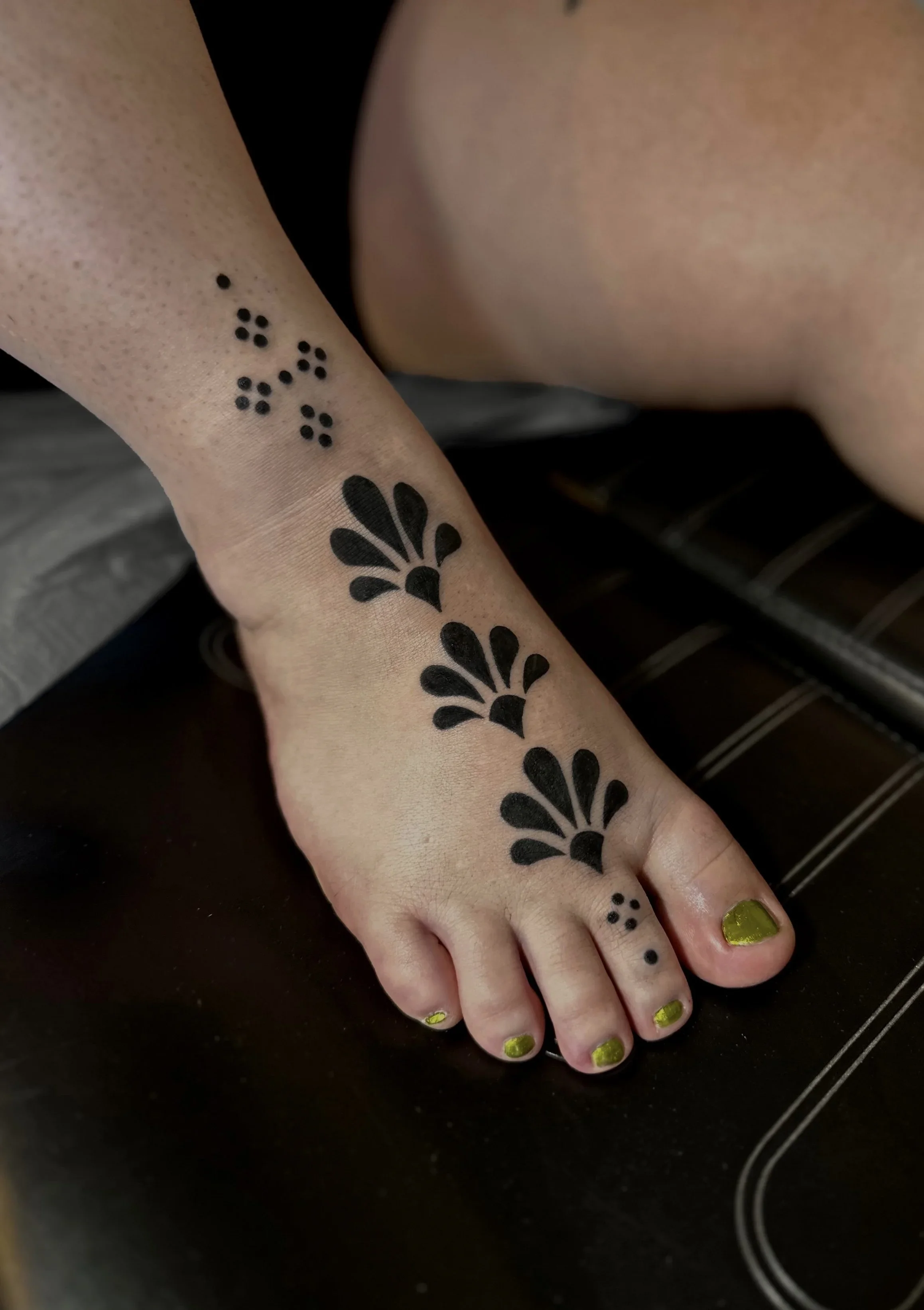 Close-up of a person's hand with black henna tattoos and yellow-green nail polish, resting on a dark surface.