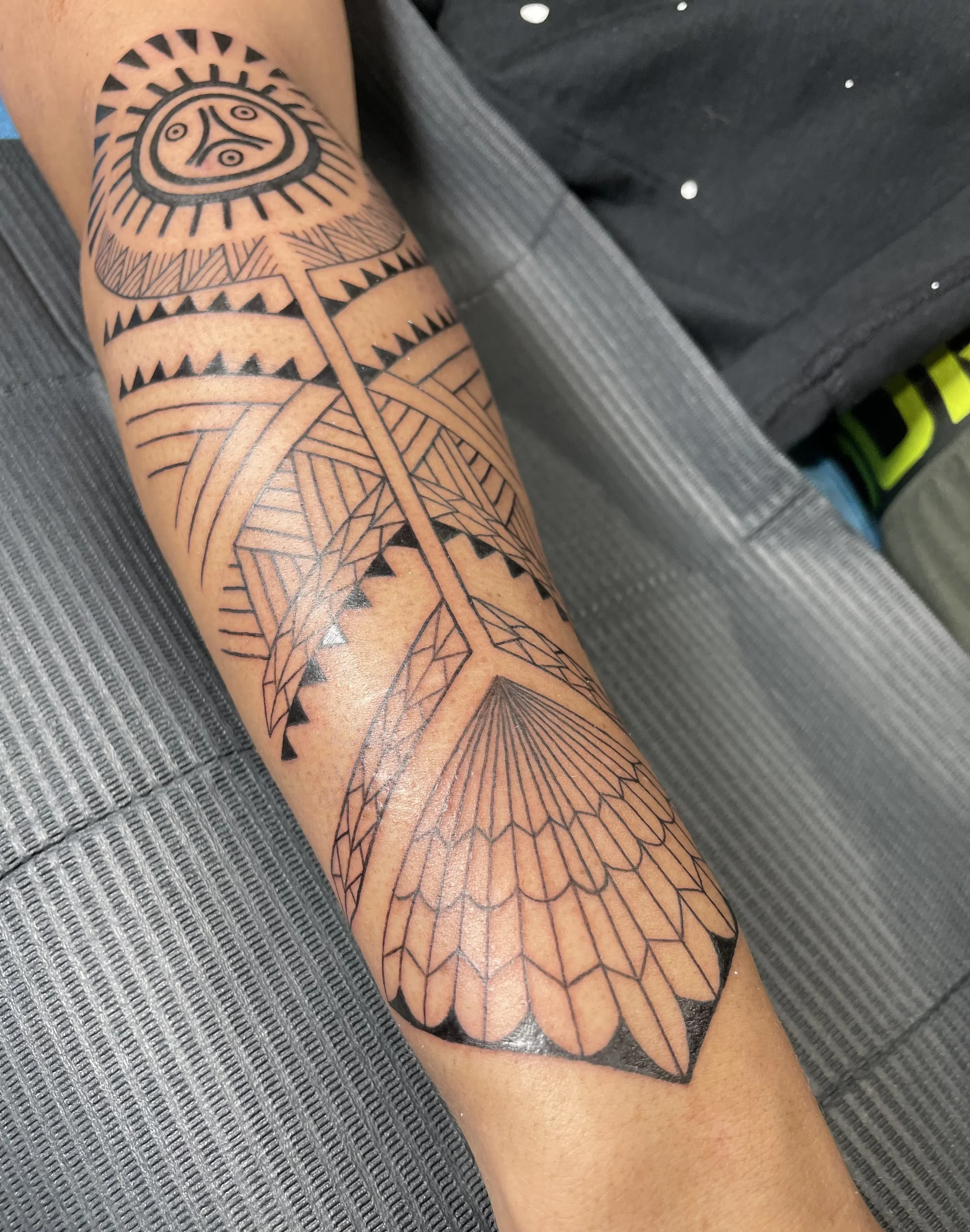 A person's arm with a large tribal-style tattoo featuring geometric patterns, lines, and shapes, including a central circular design resembling a face or mask surrounded by various patterns extending down the arm.