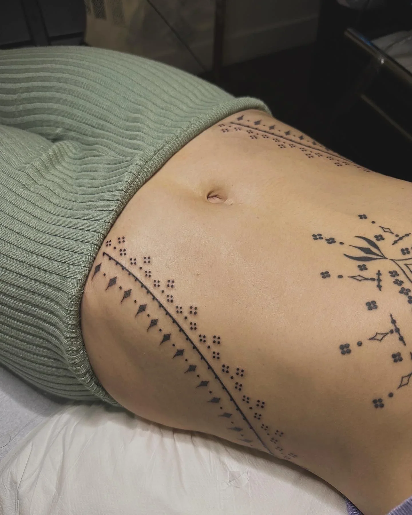 Close-up of a person's lower abdomen, showing a tattoo with floral and geometric patterns around the navel.