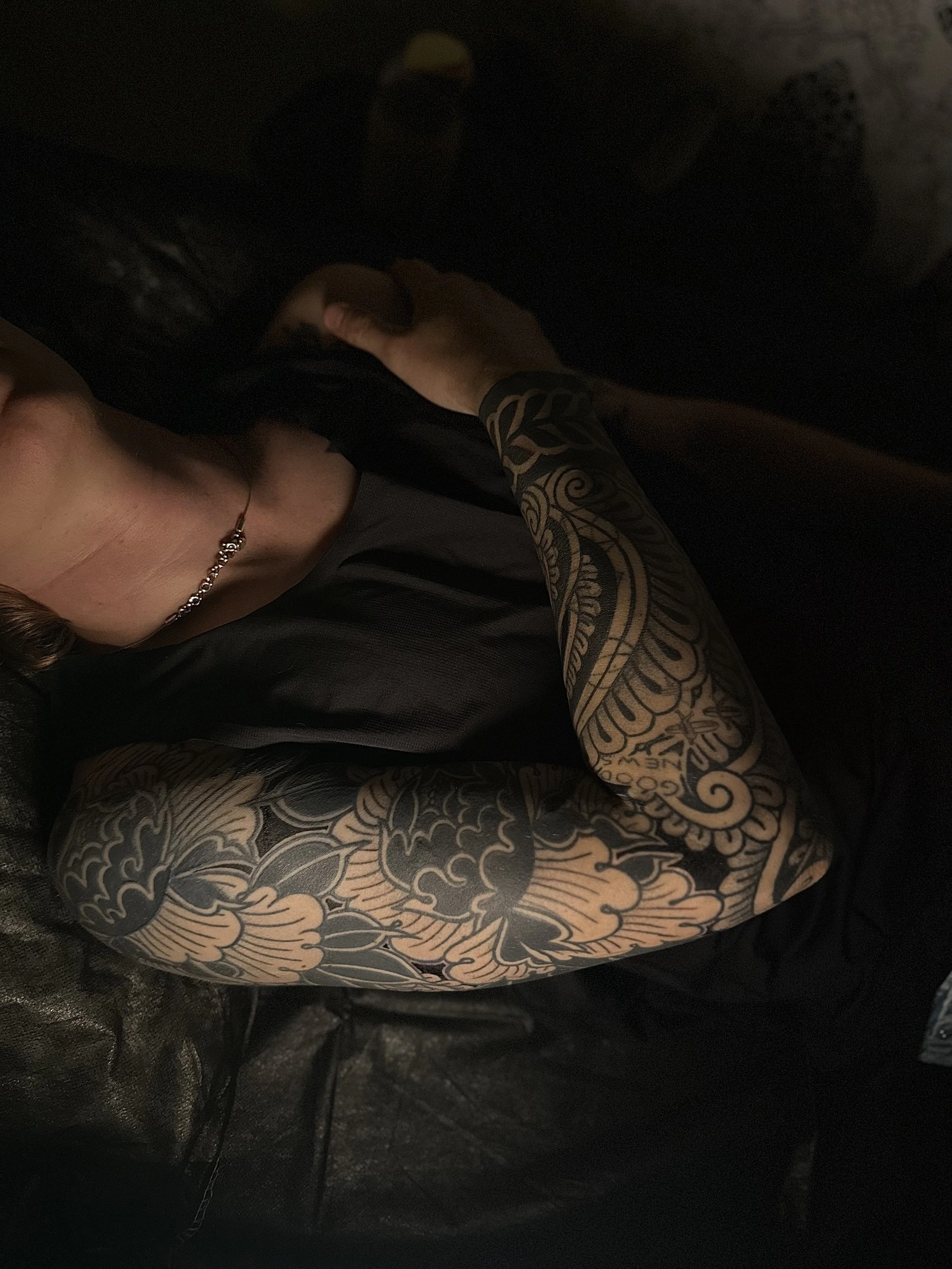 A person with a tattoo sleeve on their arm is reaching into a dark container, wearing a gray shirt, jewelry, and has long dark hair.