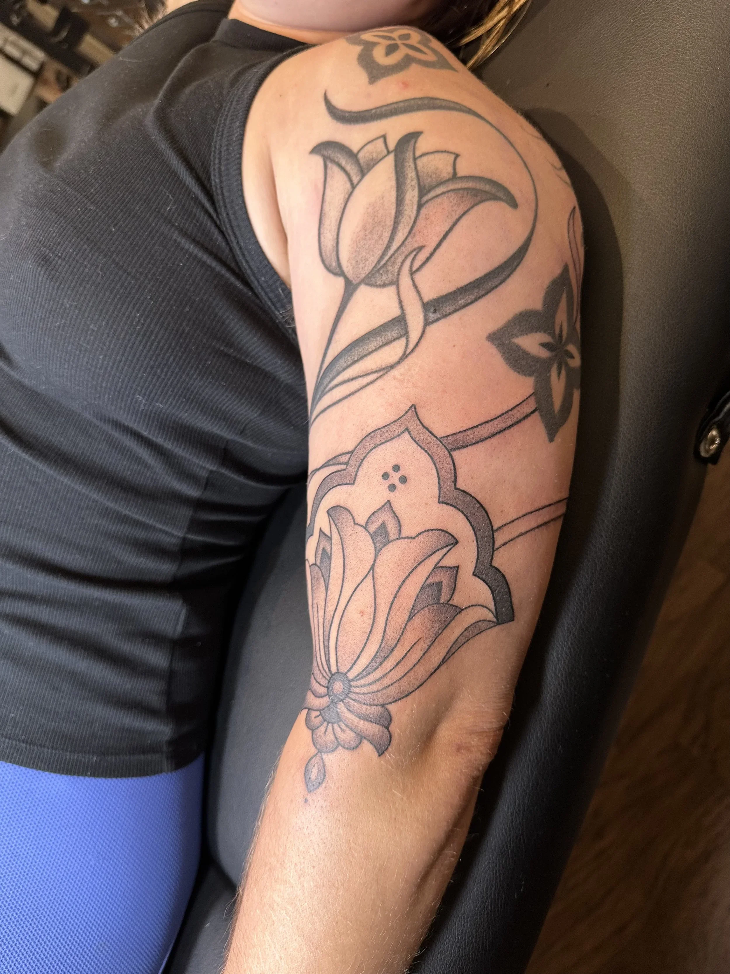 A person's upper arm with a large floral tattoo, including a prominent flower, leaves, and decorative elements, resting on a black surface.