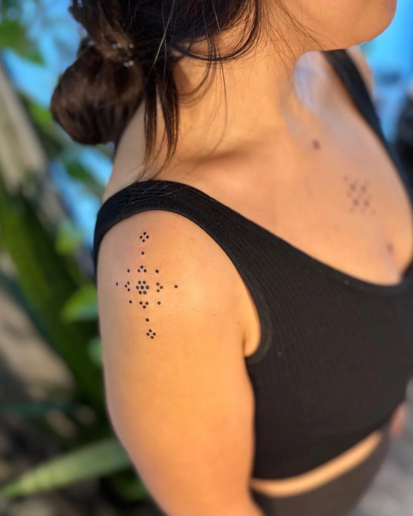 Close-up of a woman's shoulder and upper arm, showing black dot tattoos arranged in geometric patterns on her upper arm and shoulder. She is wearing a black sleeveless top, and her hair is dark and wavy, partially covering her face. The background is