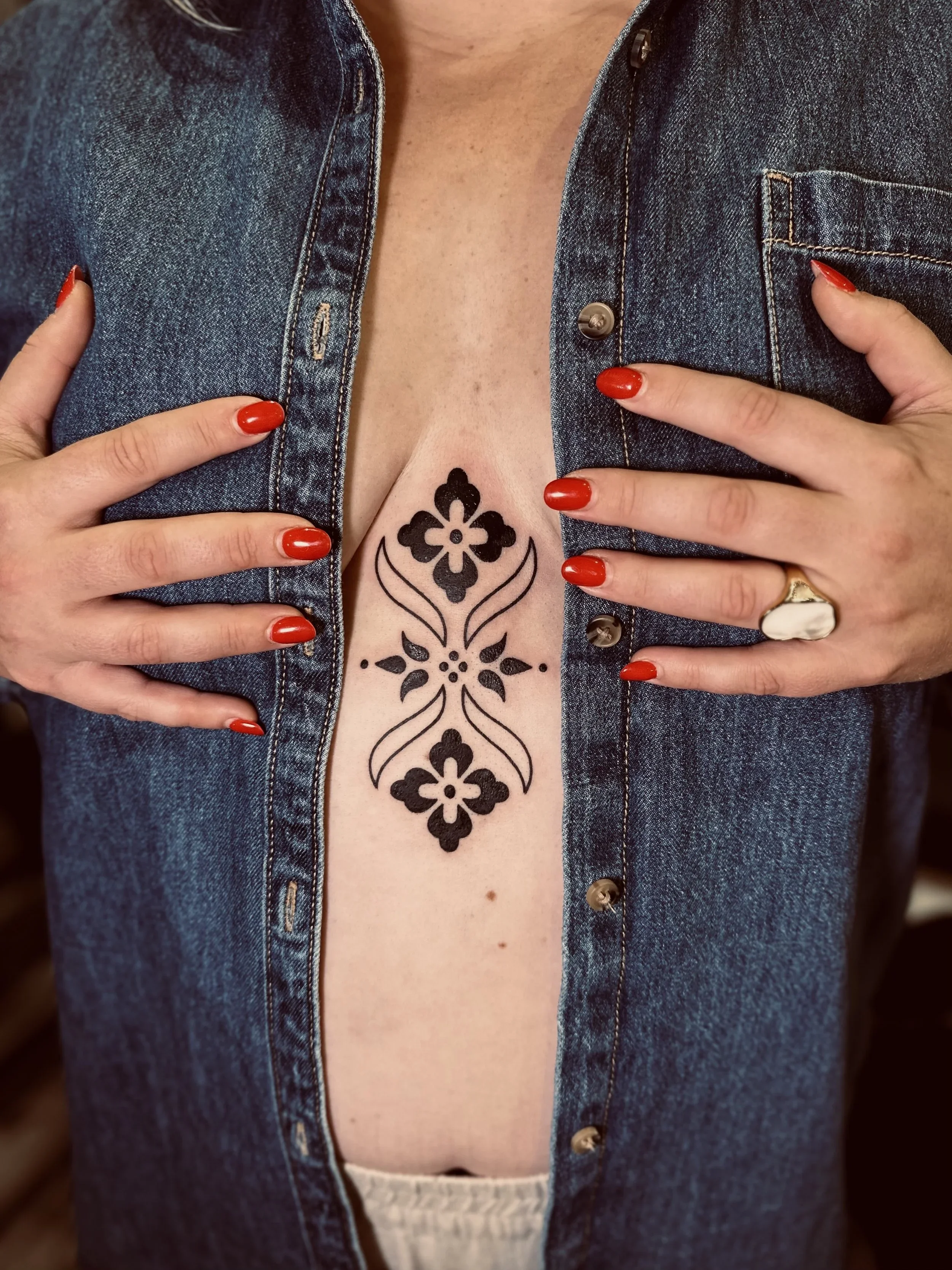 Close-up of a person wearing a denim jacket with hands on chest, red nail polish, and a black floral tattoo visible on the chest.